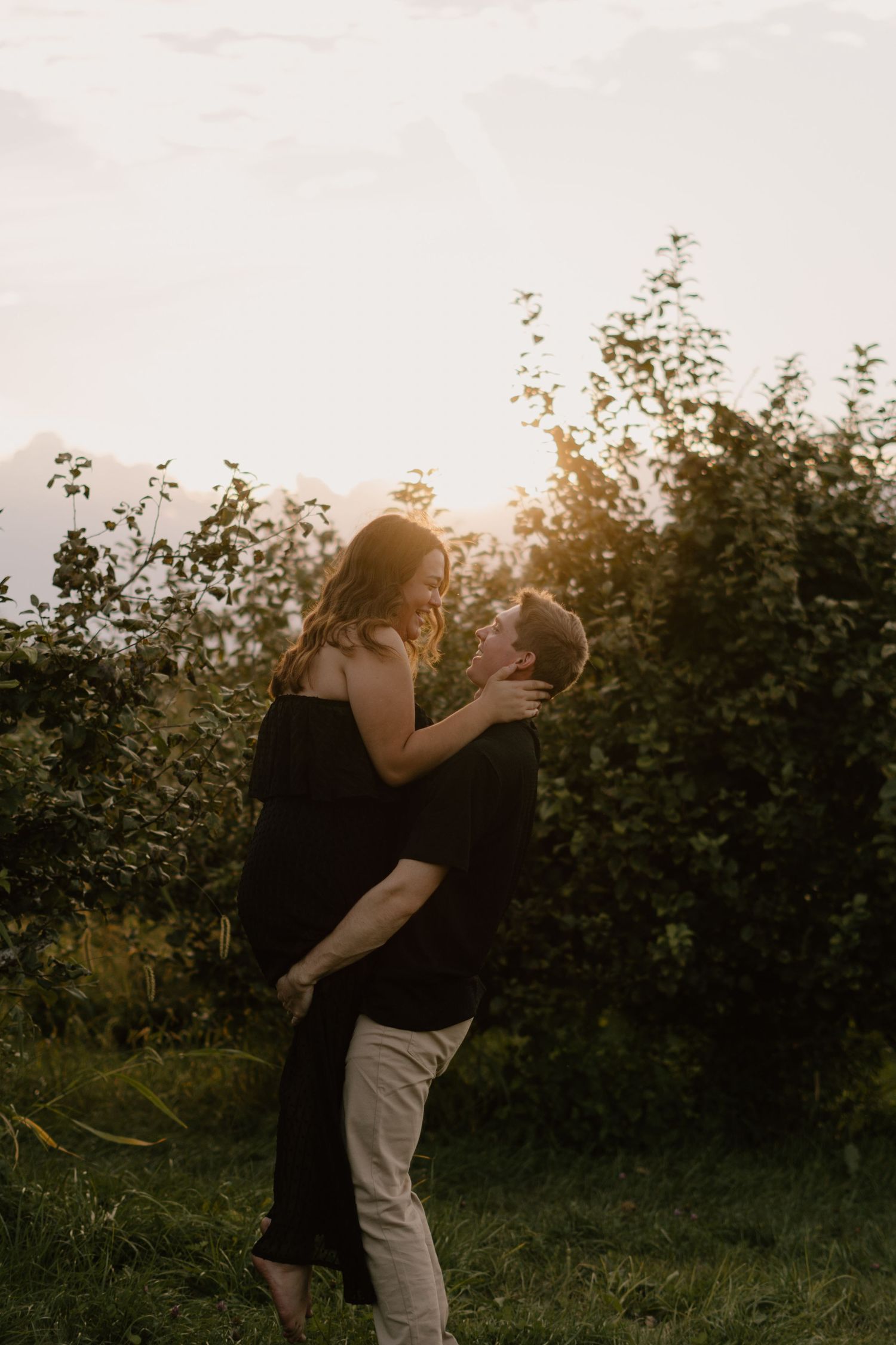 Couple embracing at golden hour in lush green orchard, man lifting woman in black dress, romantic sunset backdrop.