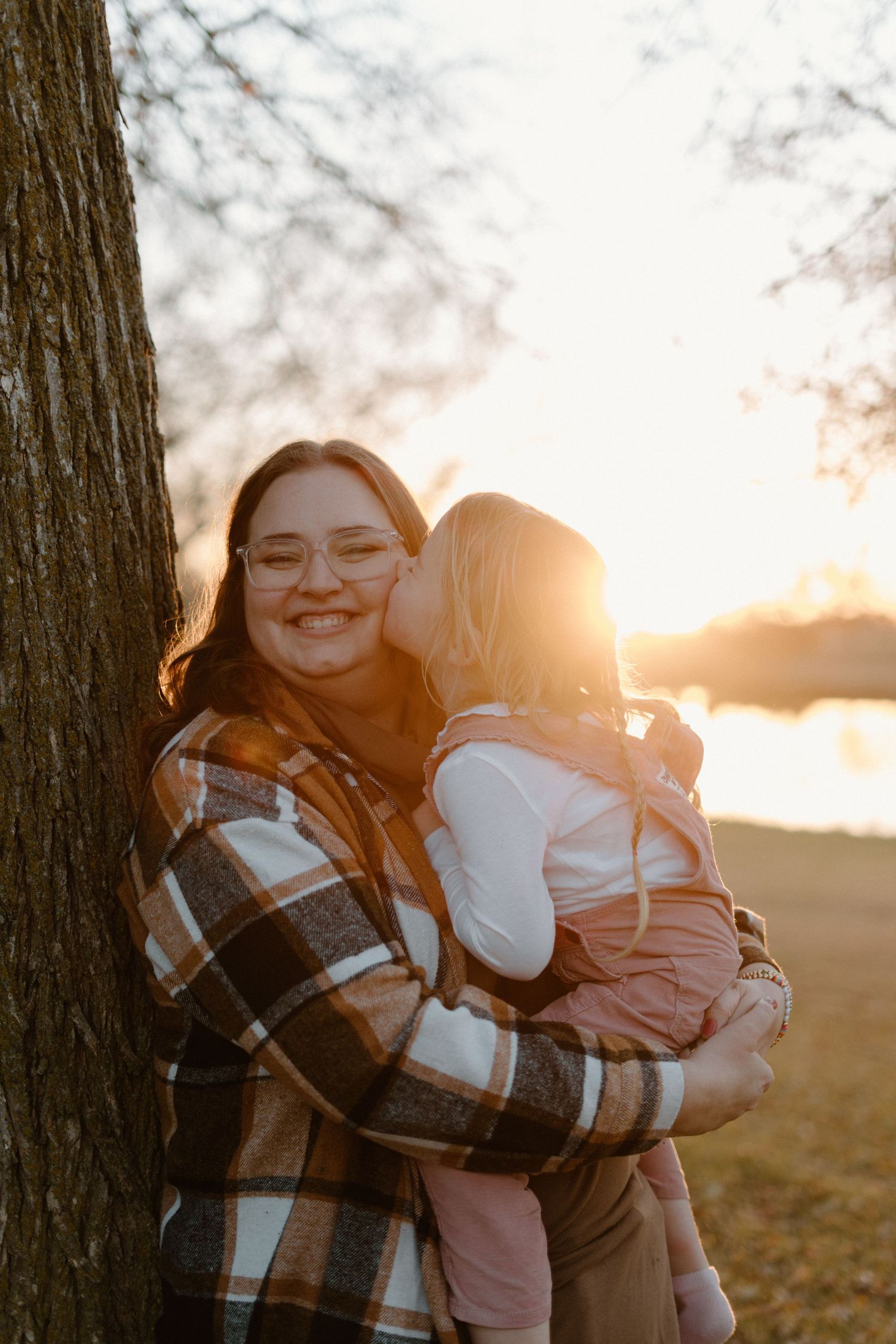 Smiling woman with glasses holding a child who kisses her cheek at golden sunset.