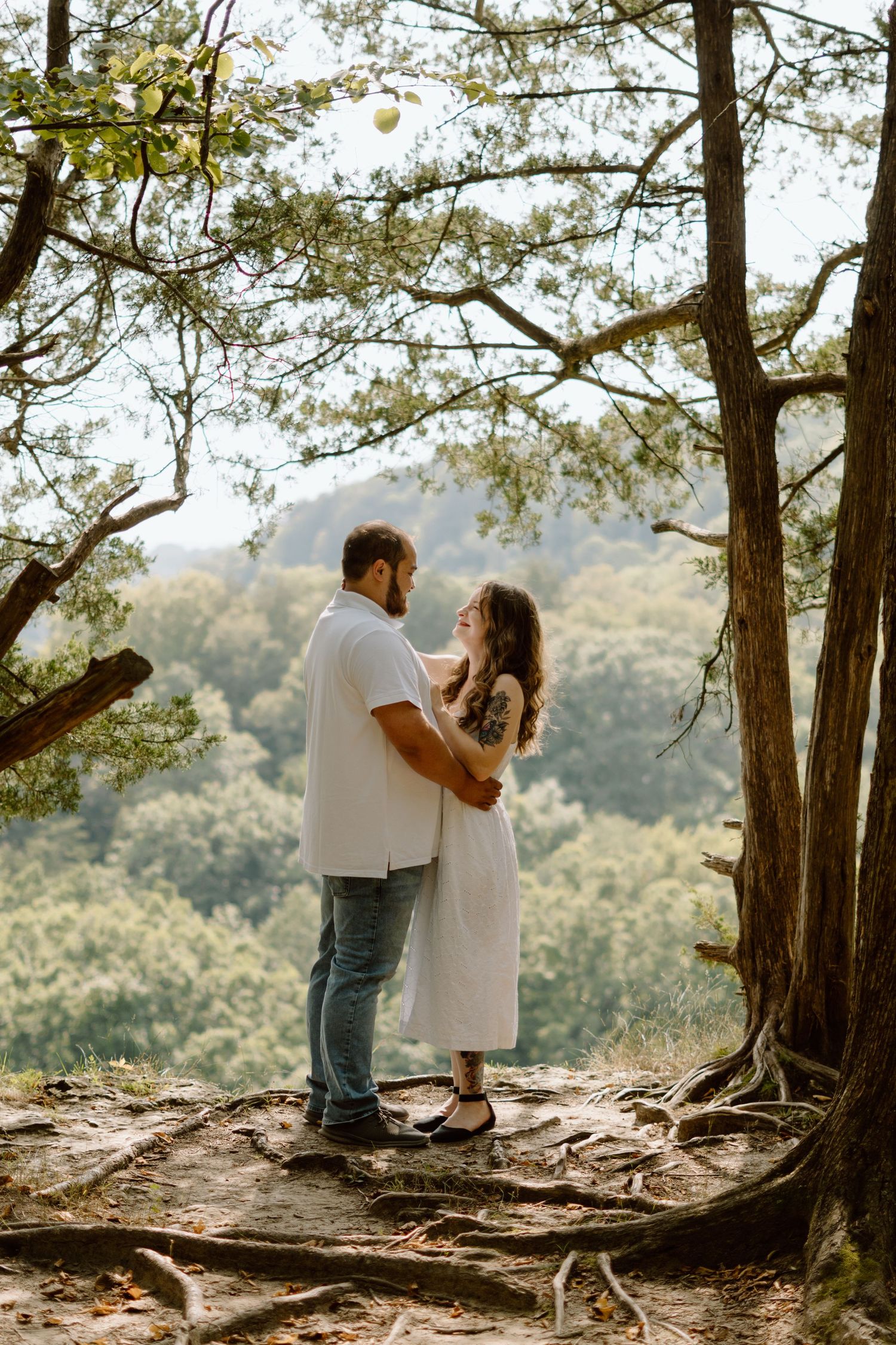 A couple embraces romantically under a large tree overlooking a lush green mountain landscape.
