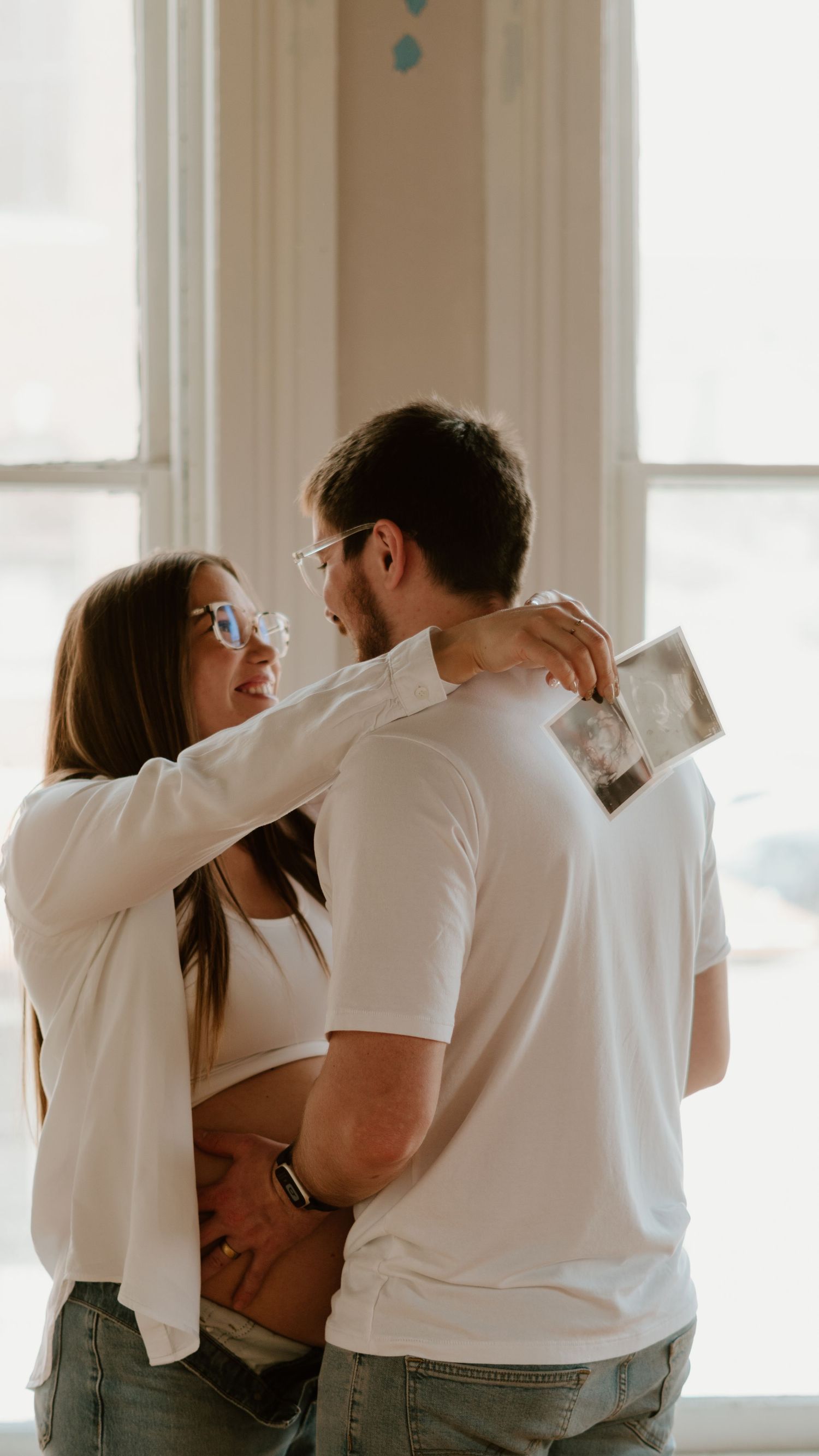 Expecting couple in white outfits holding ultrasound photo while embracing near a bright window indoors.
