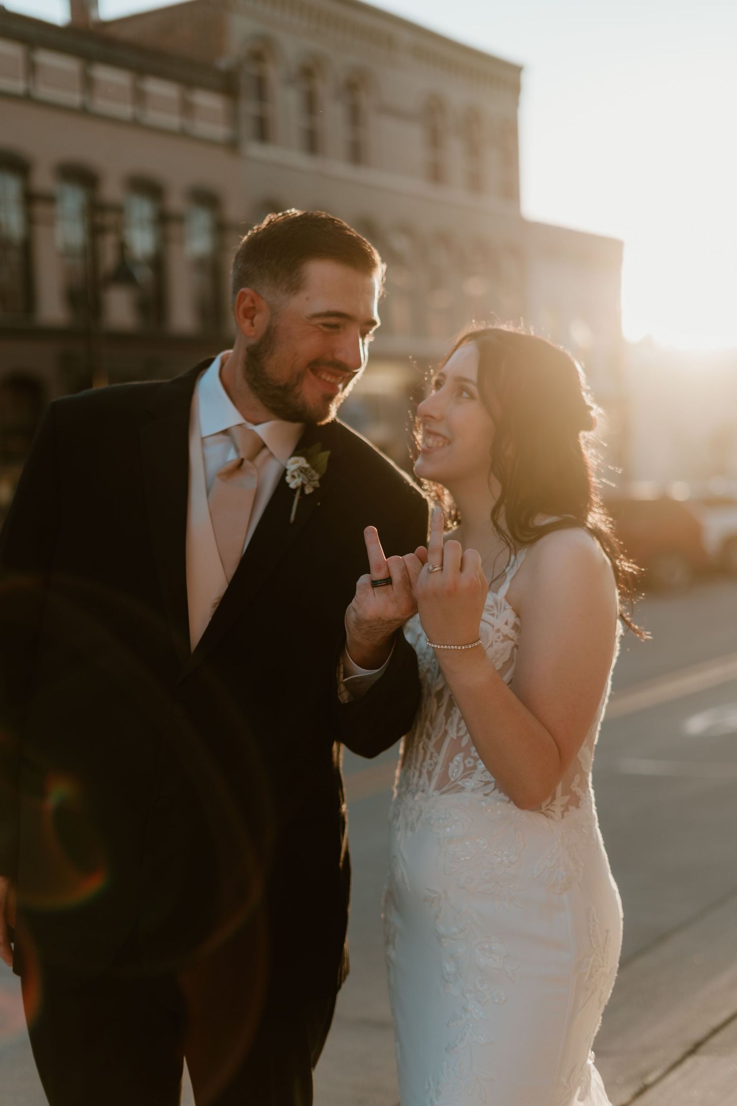 Bride and groom share a joyful moment at golden hour on a city street, bride flashing a shaka sign.