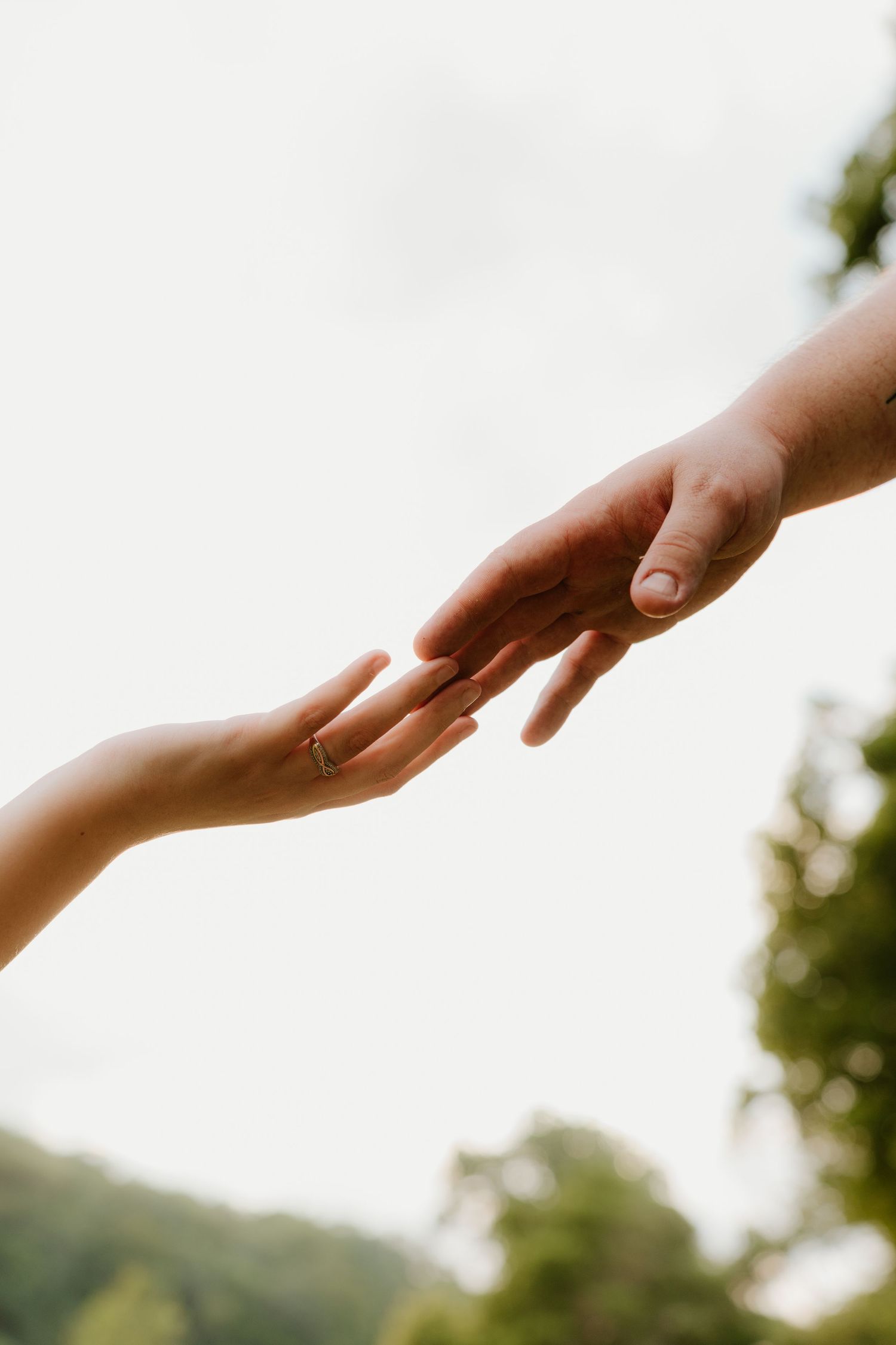 Two hands reaching toward each other outdoors, one wearing a ring, against a soft green and white background.