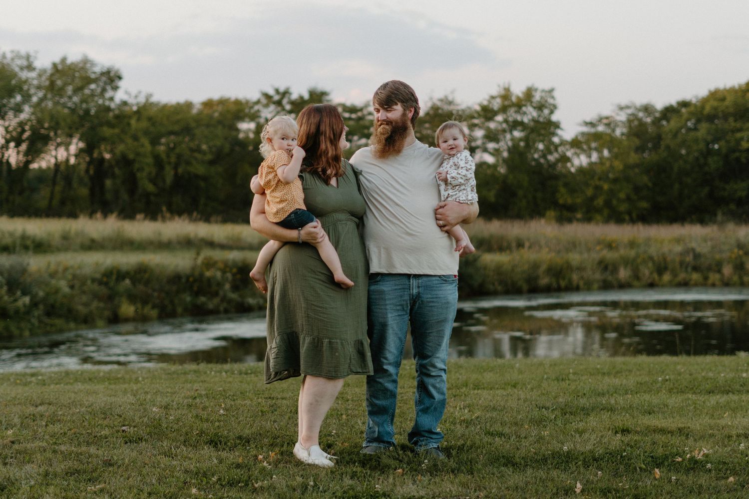 Family of four posing outdoors near a pond, parents holding two toddlers in a lush green meadow at dusk.