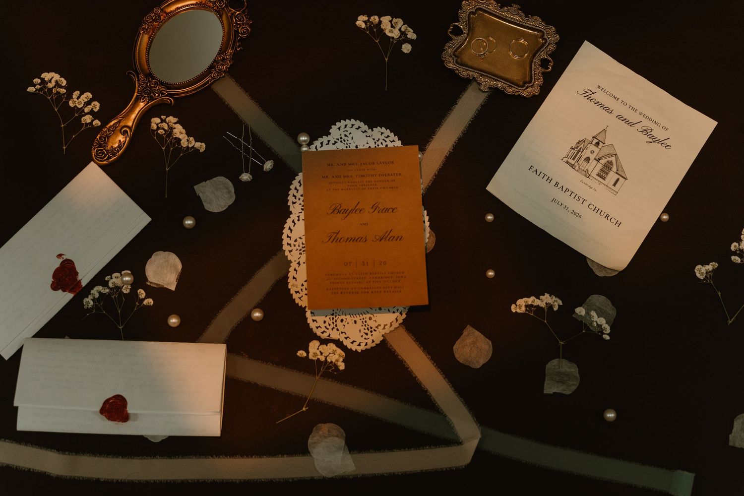 Elegant wedding stationery flat lay with wooden invitation, doily, envelopes, and floral accents on dark background.