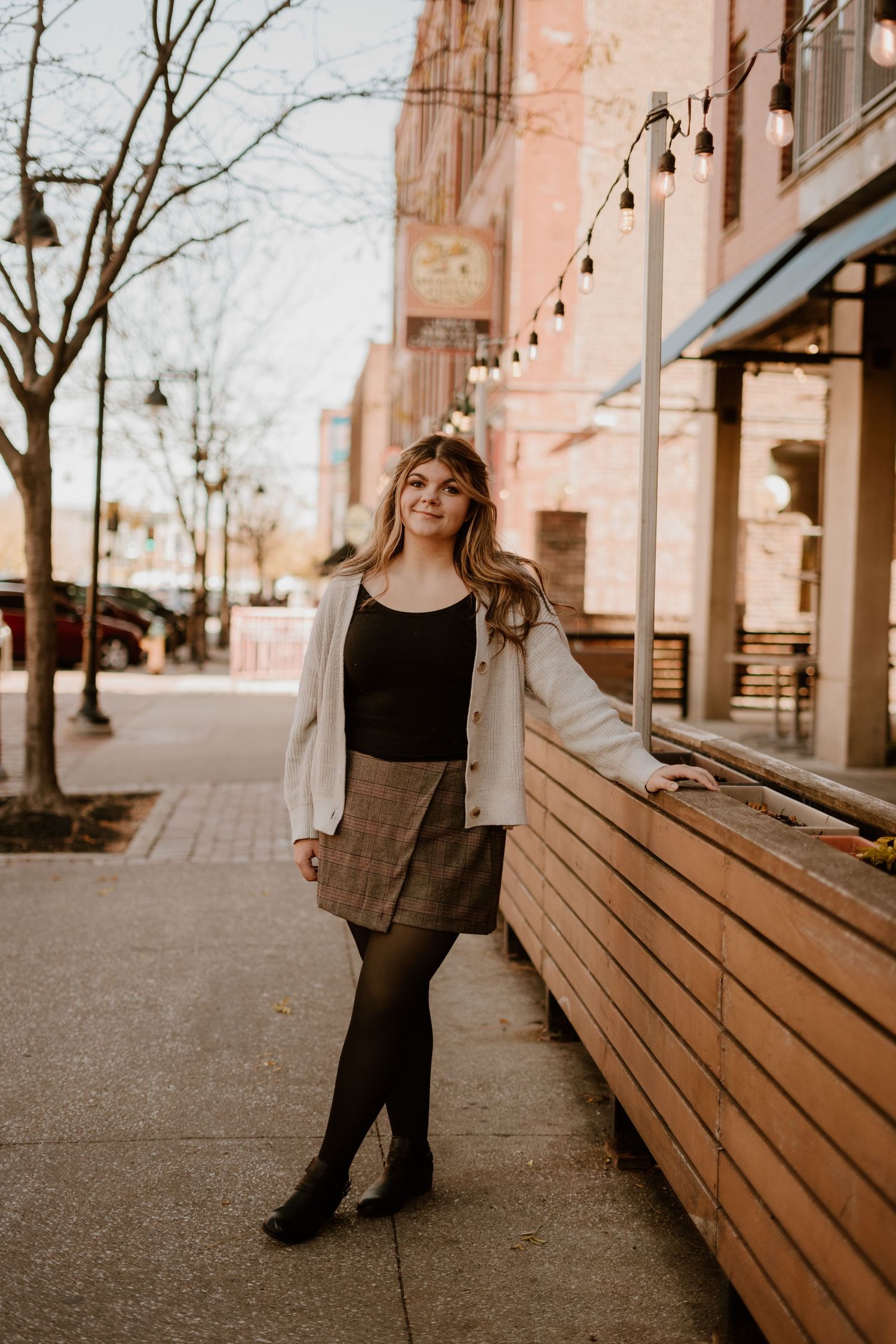 Young woman in plaid skirt, black top, and white jacket posing on urban sidewalk with string lights and brick buildings.