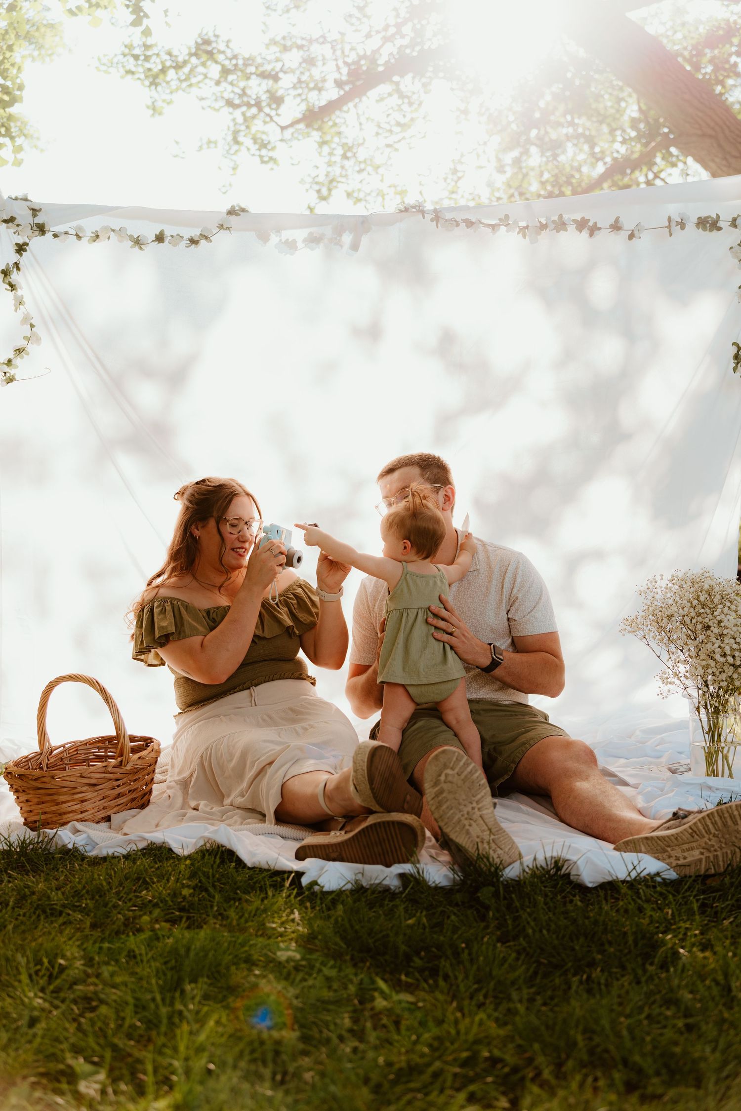 Family enjoying a sunny outdoor picnic on a white blanket with a baby, wicker basket, and floral backdrop.