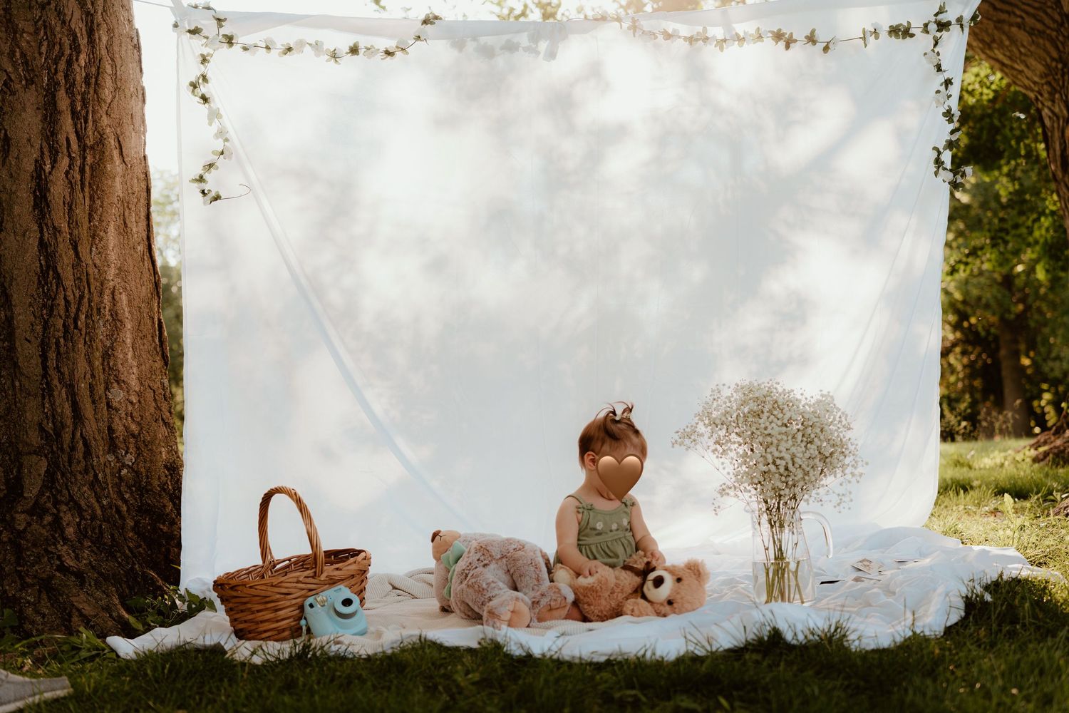 A baby sits on a white blanket outdoors with stuffed animals, a basket, and flowers against a white backdrop.