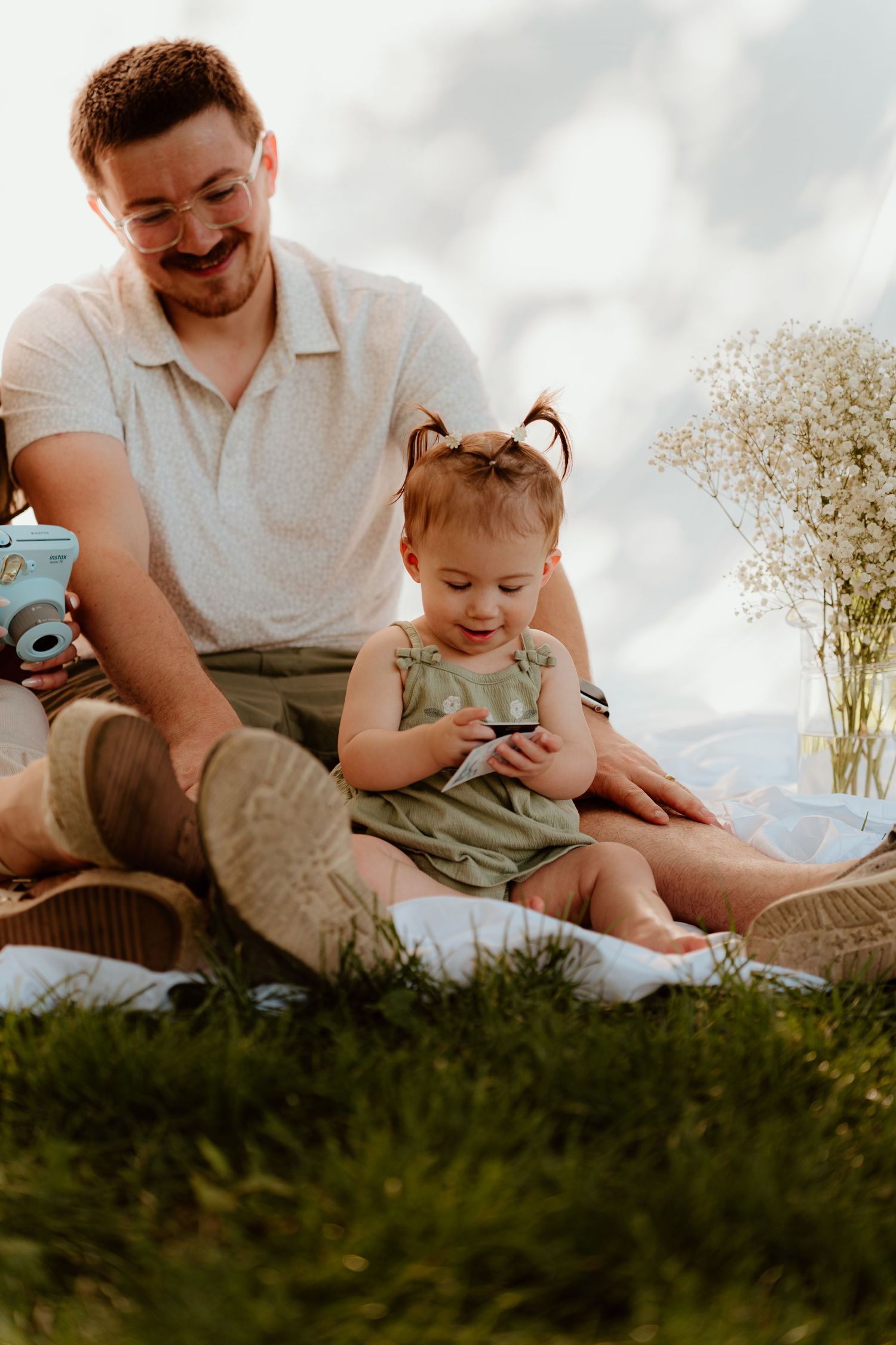 Father and toddler daughter enjoying outdoor picnic on grass with flowers and vintage camera.