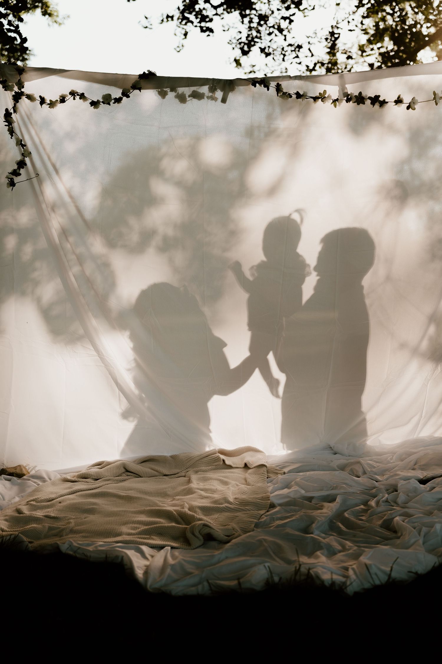 Silhouettes of two adults holding a baby projected on a white fabric backdrop outdoors.