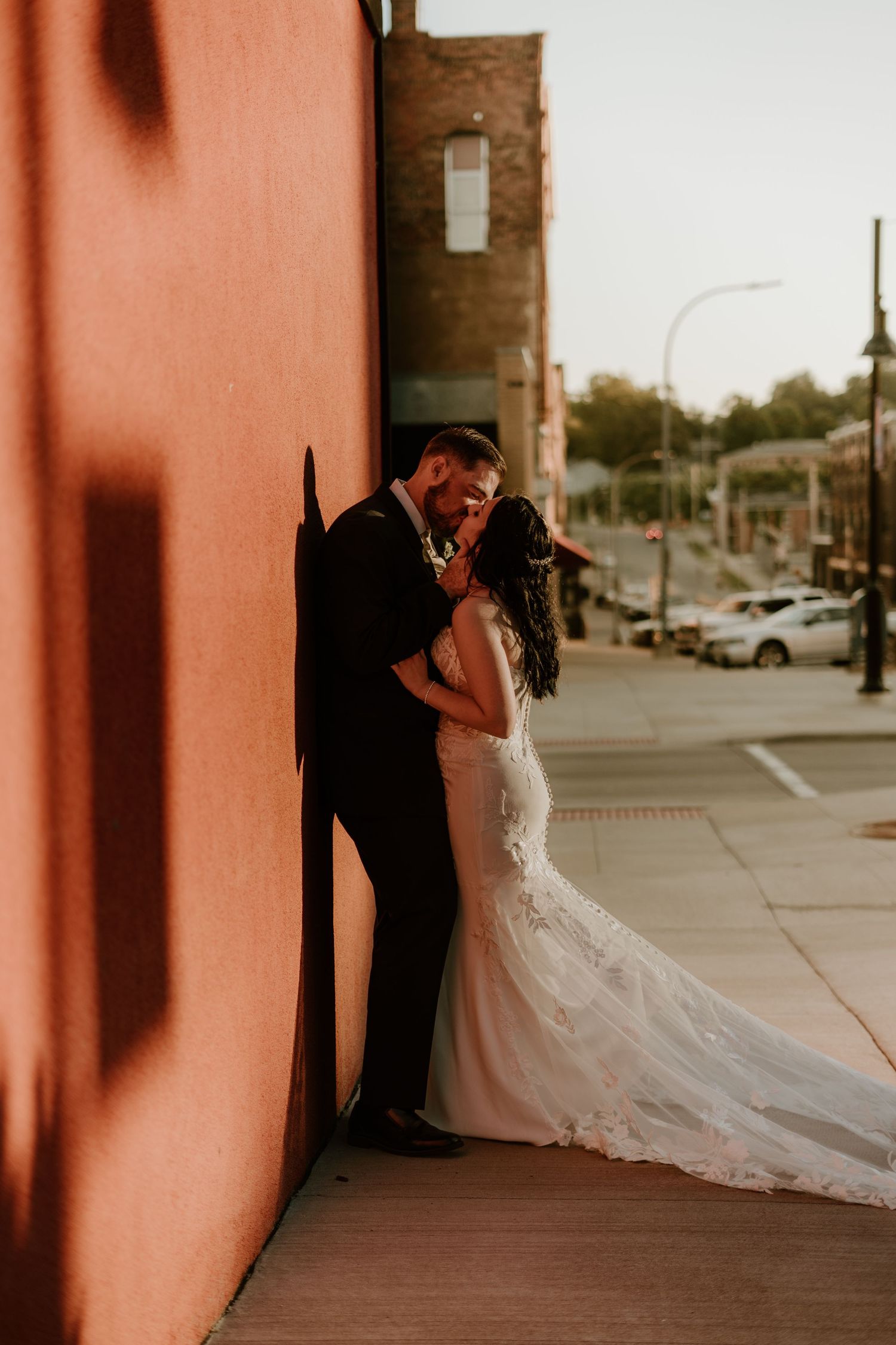 Bride and groom share a romantic kiss against a rustic red wall during golden hour in an urban setting.