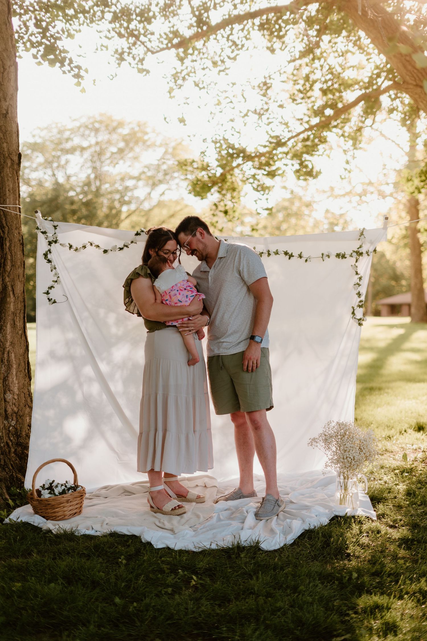 Couple sharing a tender moment during an outdoor maternity photo shoot with a white backdrop under a sunlit tree.
