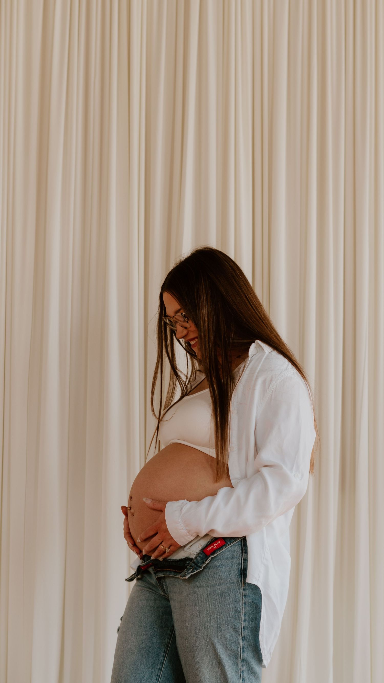 A pregnant mama in a white shirt and jeans cradles her baby bump while standing before white curtains.