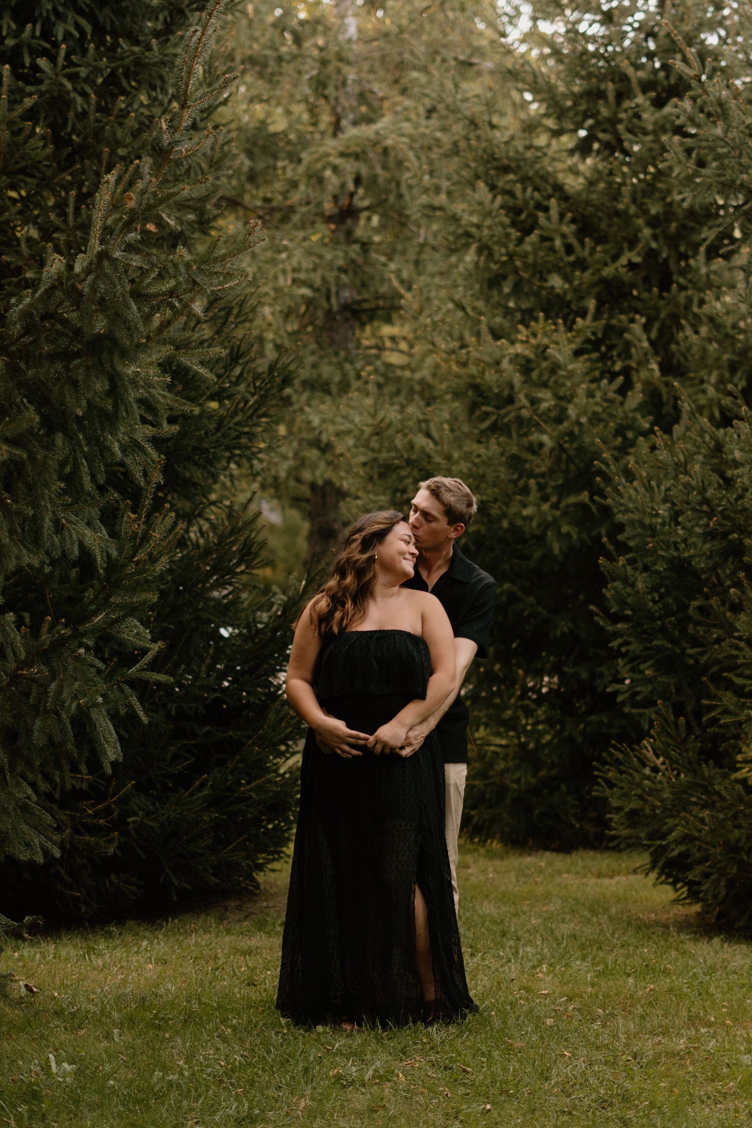 Couple embracing in black outfits among lush evergreen trees, romantic outdoor engagement photo session.