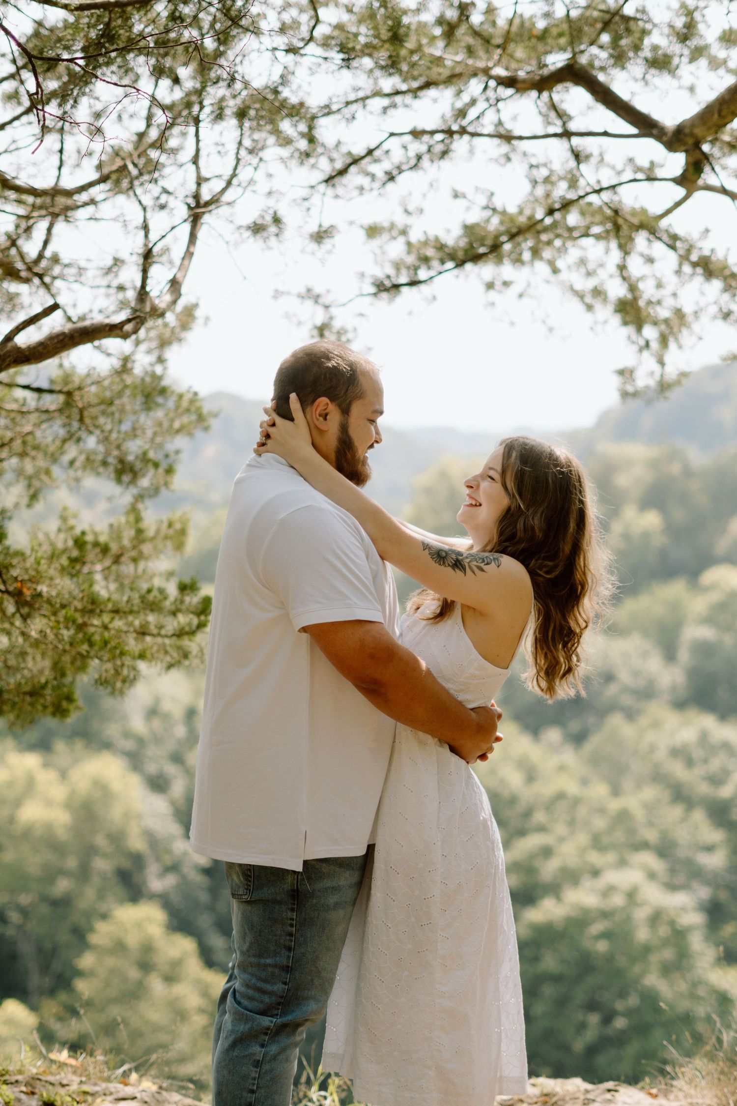 Happy couple embraces and smiles at each other outdoors under a tree with lush green mountain views behind them.