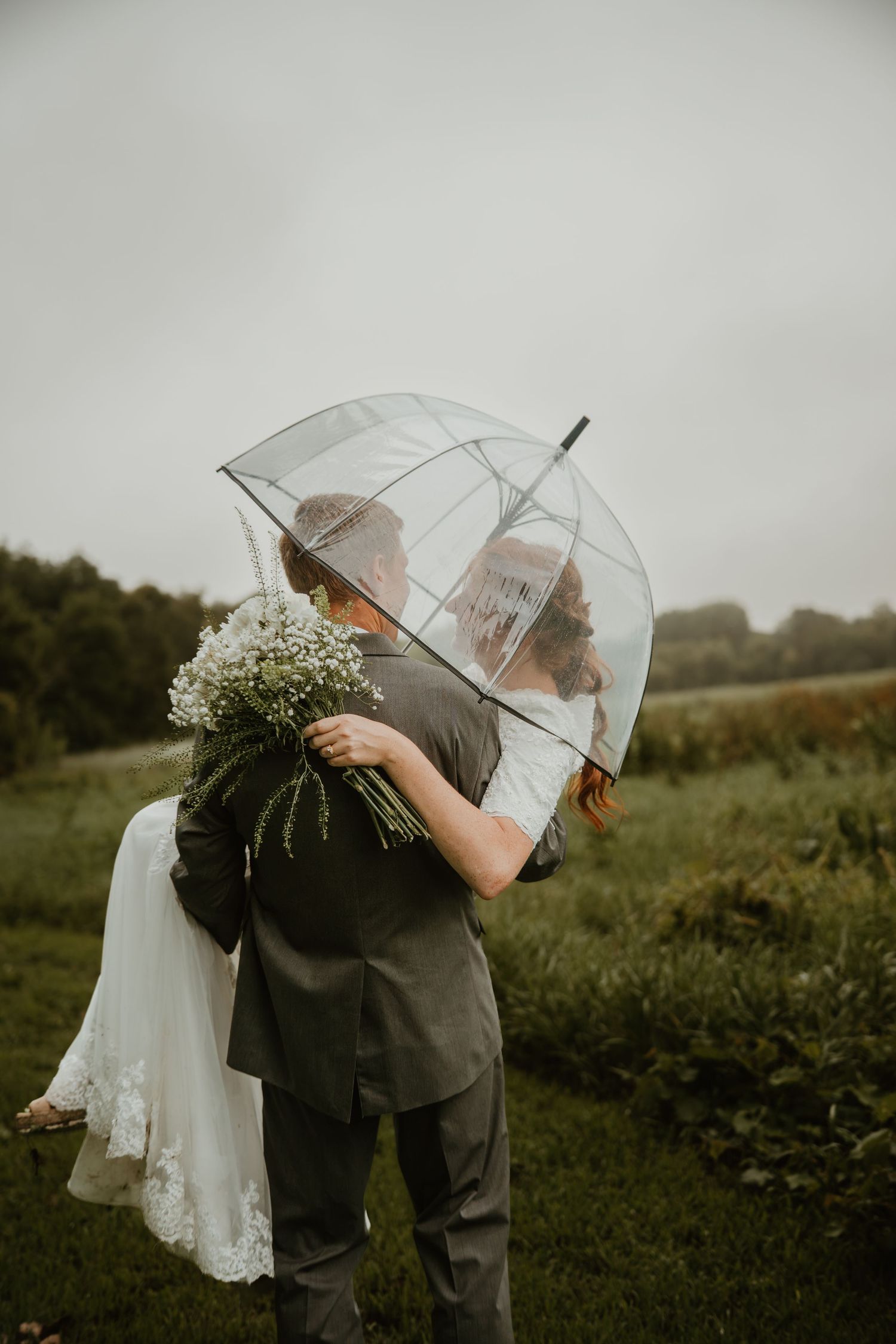 Bride and groom kissing under a clear bubble umbrella in a green field, bride holding a white wildflower bouquet.