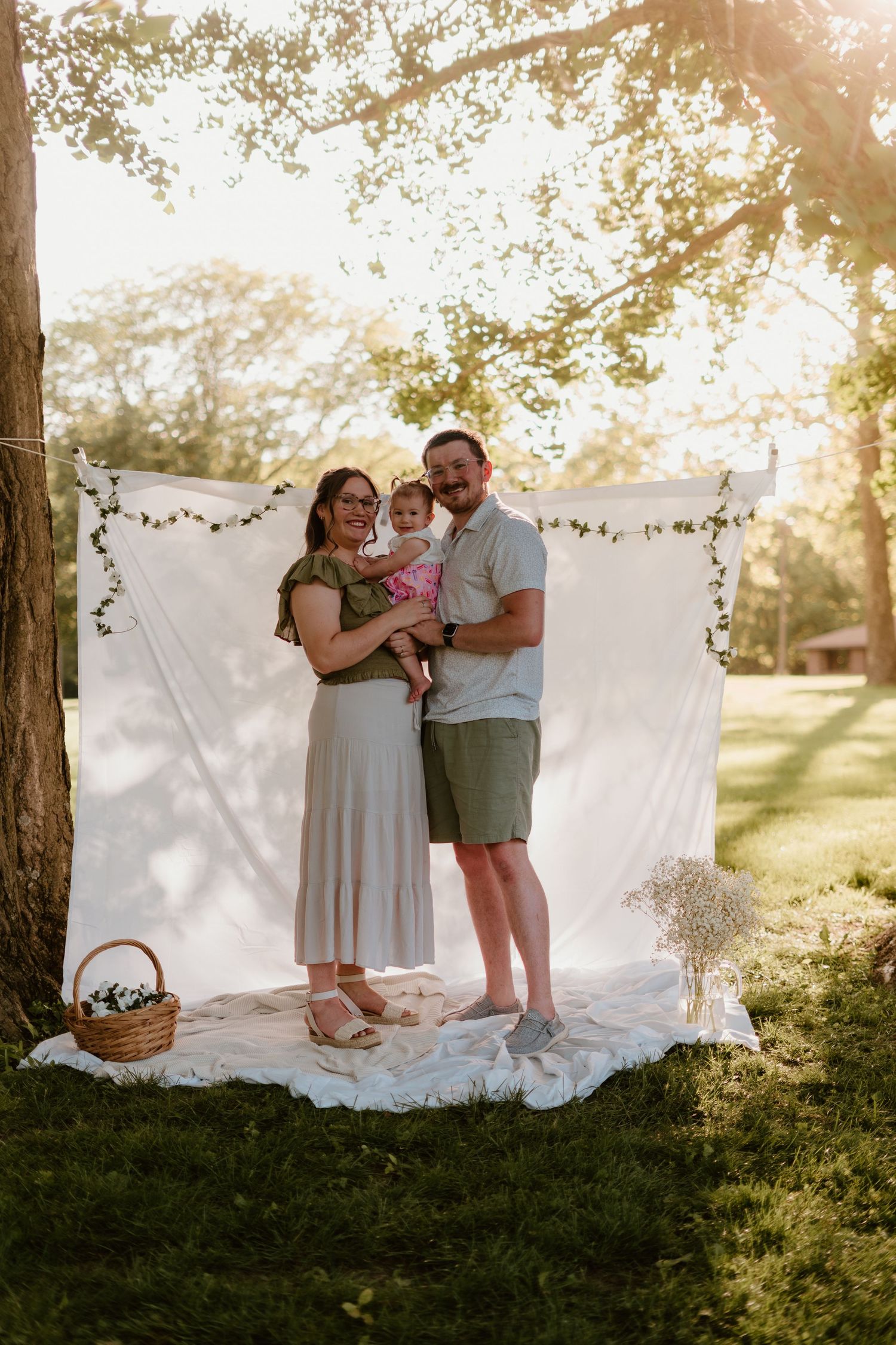 A young family with a baby poses outdoors in front of a white fabric backdrop with greenery at golden hour.