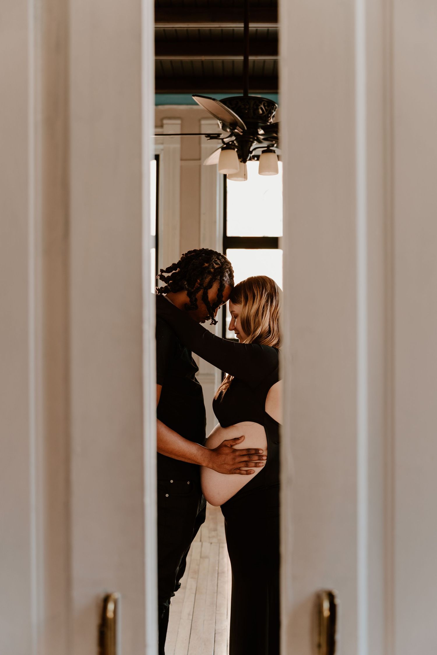 Couple sharing tender moment in doorway, hands cradling pregnant belly, wearing matching black outfits.