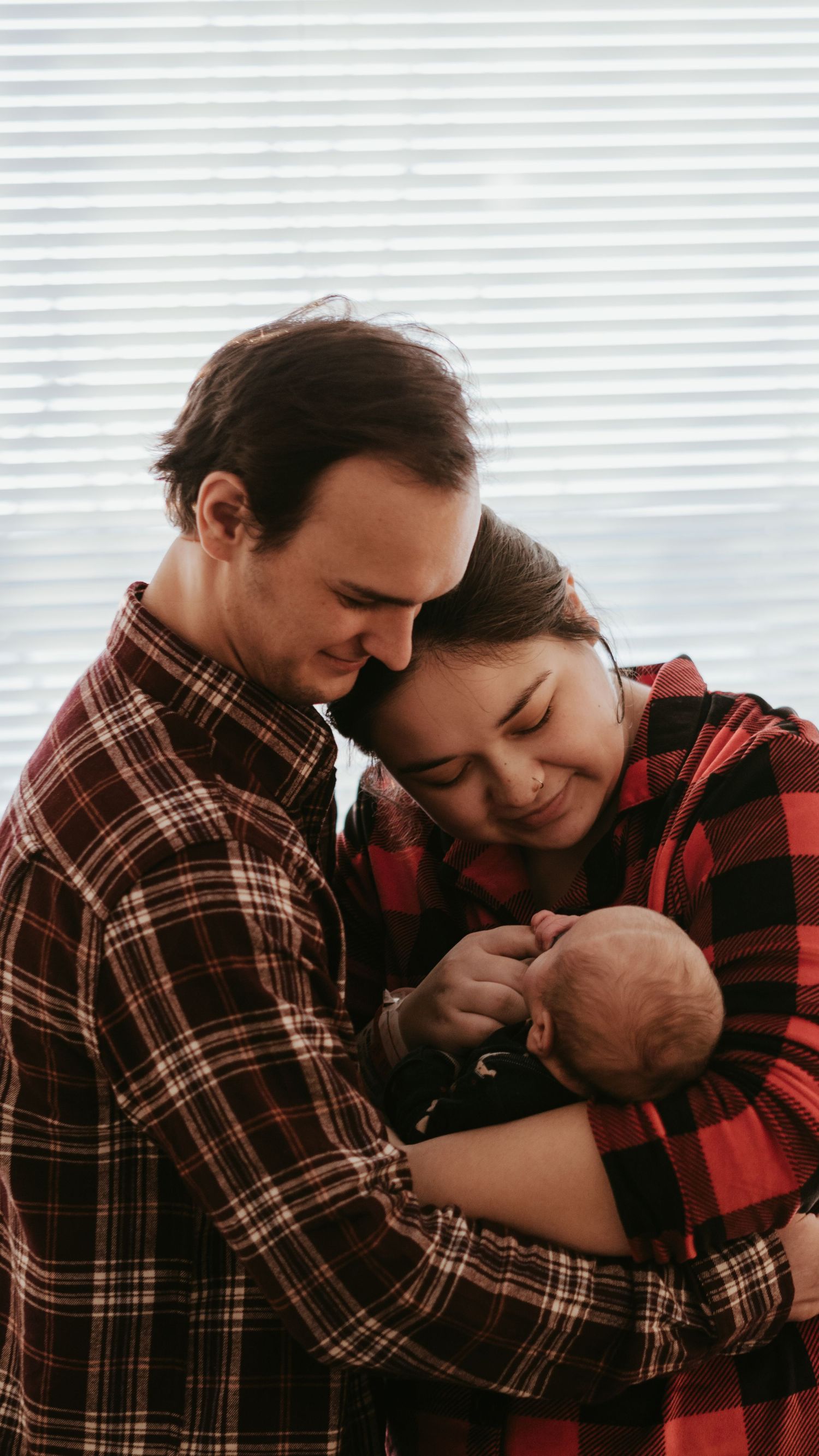 New parents in matching red plaid shirts tenderly hold their newborn baby together in a warm family portrait.
