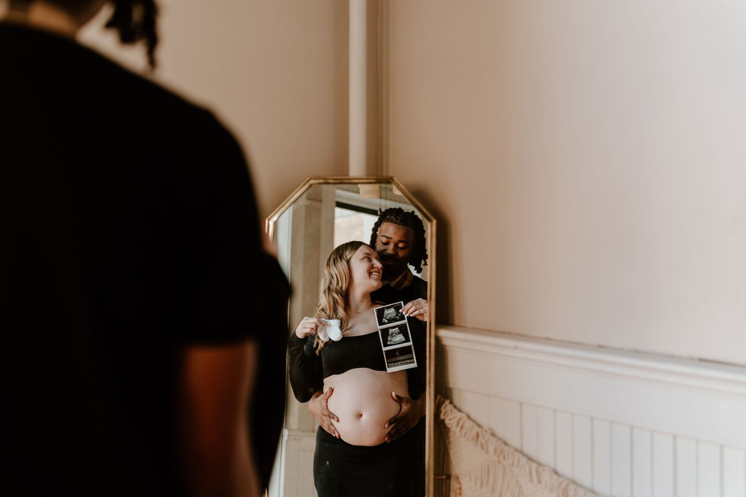 Couple sharing a tender moment in mirror reflection, woman showing ultrasound photo during maternity photoshoot.