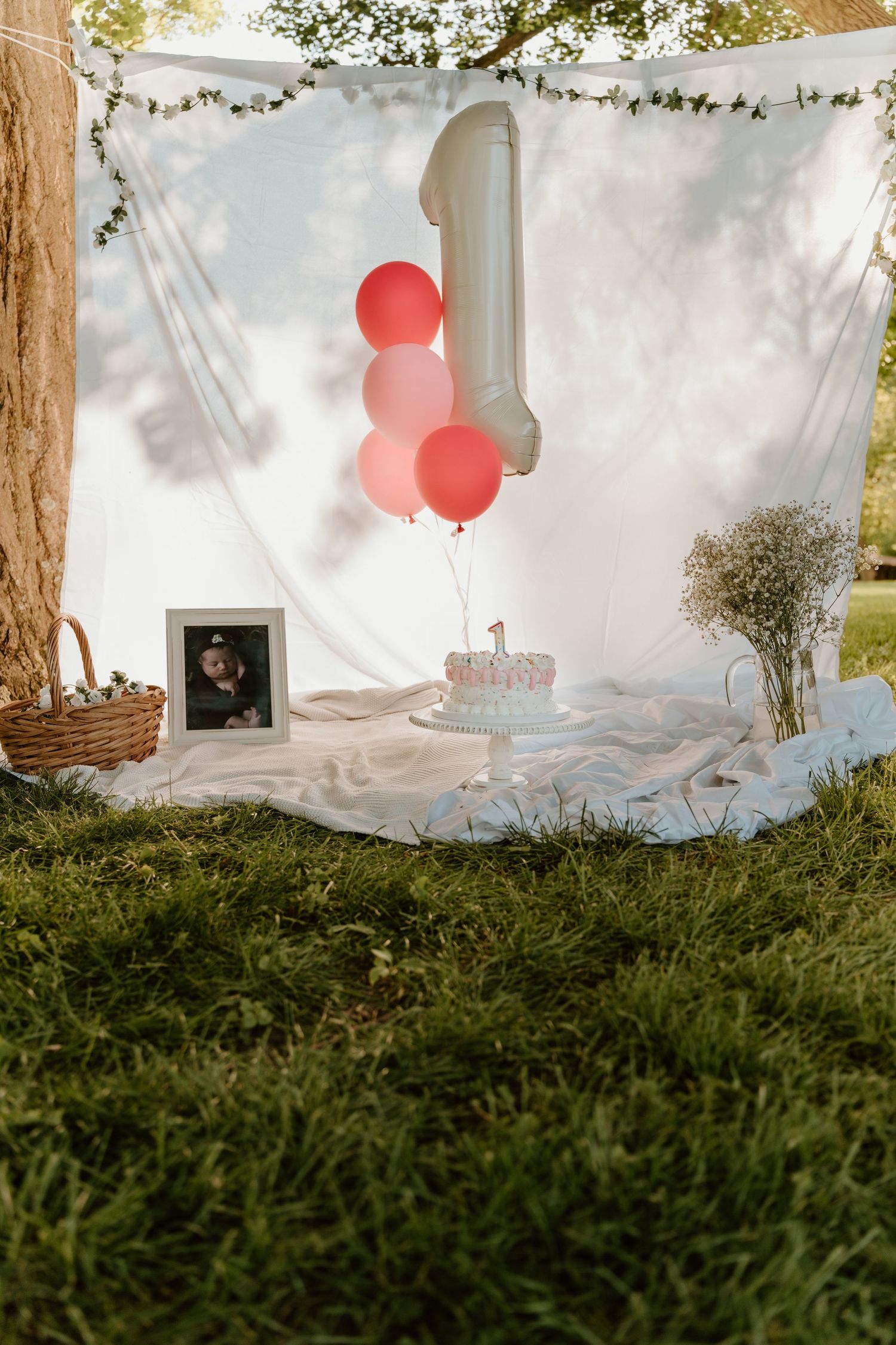 Outdoor first birthday setup with pink balloons, number 1 balloon, cake, and framed photo on white blanket.