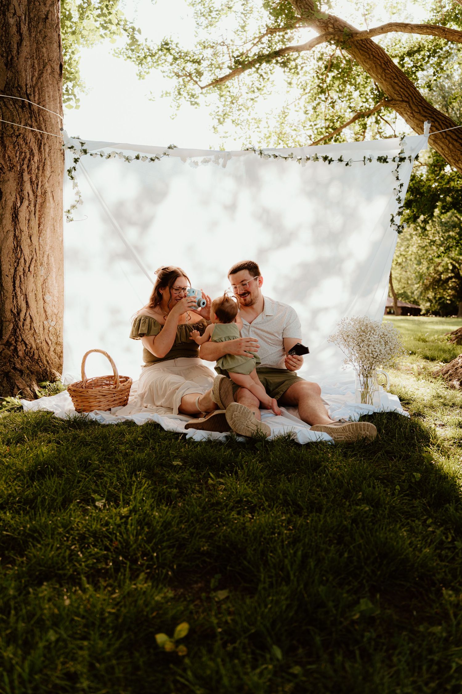 Family enjoying a sunny outdoor picnic on a white blanket under a tree with a white backdrop and string lights.
