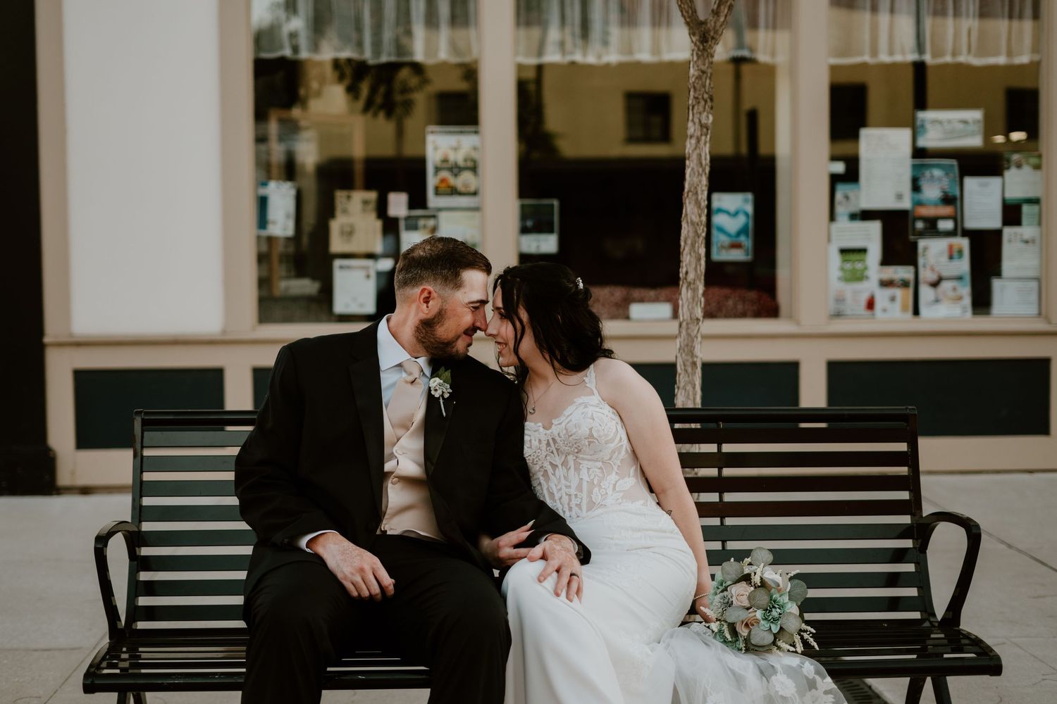 A bride and groom sit closely on a bench outside, sharing an intimate moment in wedding attire.