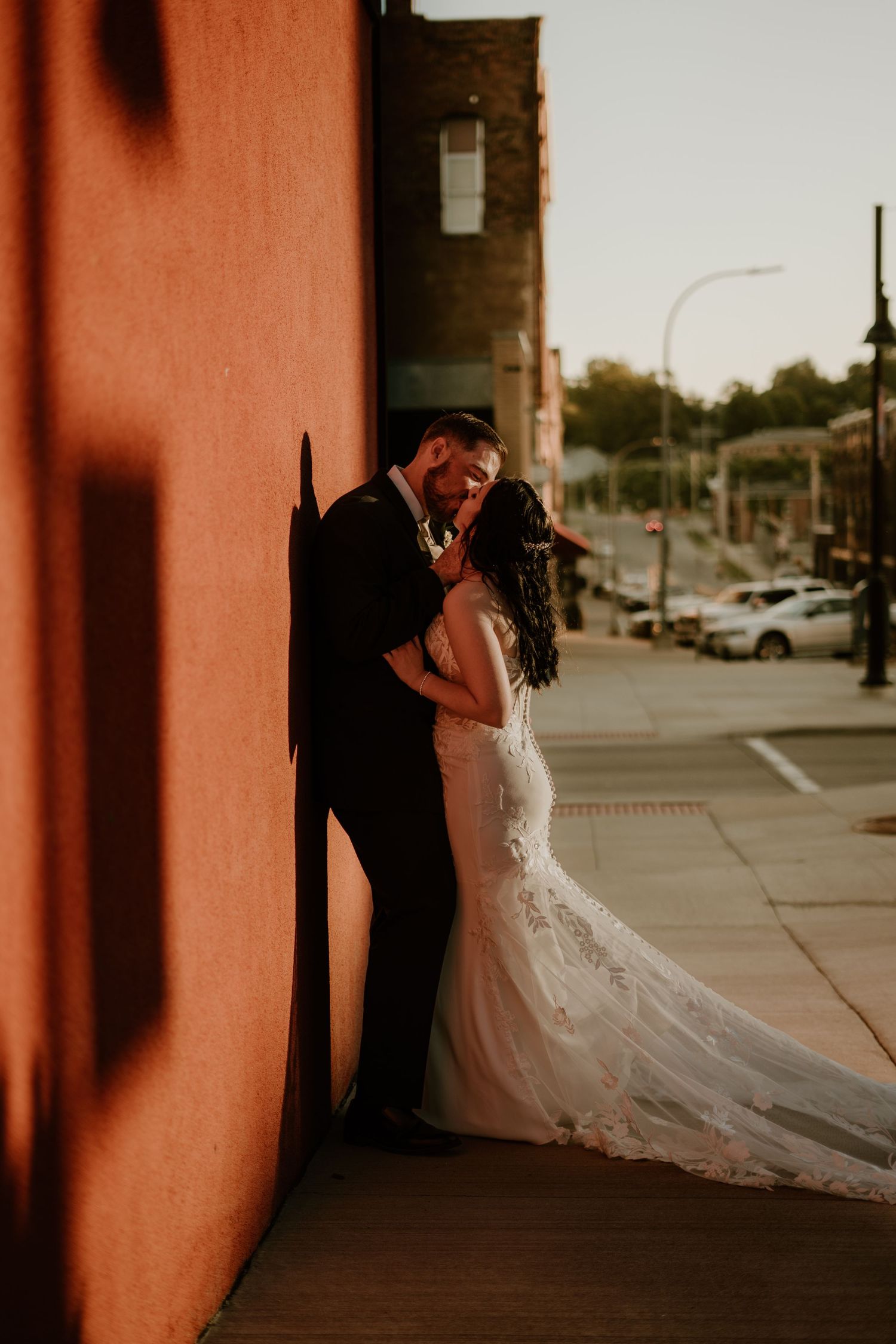 Bride and groom share a romantic moment leaning against a rustic red wall at golden hour on a city sidewalk.