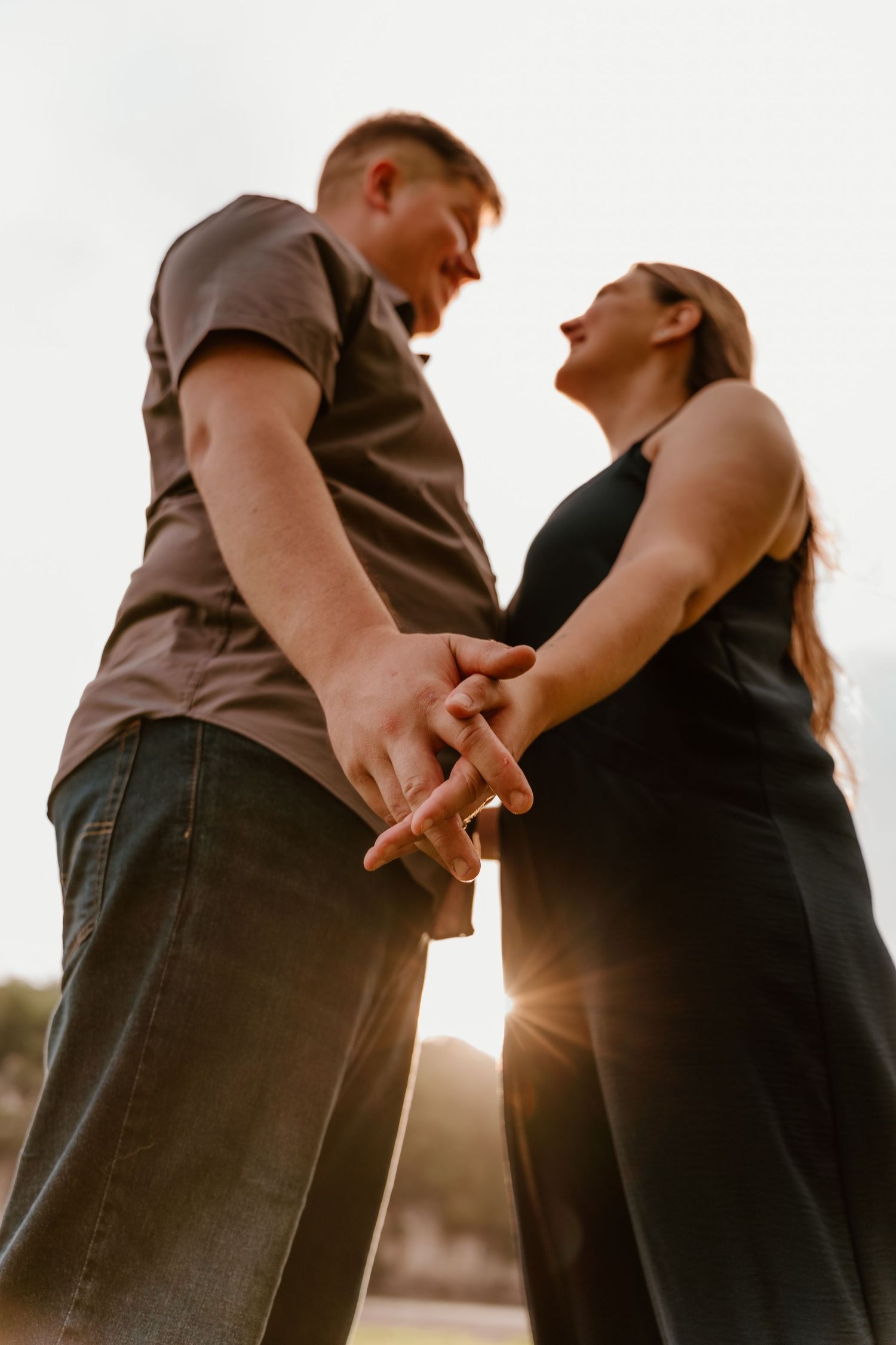 Couple holding hands at sunset, smiling at each other, man in brown shirt and woman in black dress outdoors.