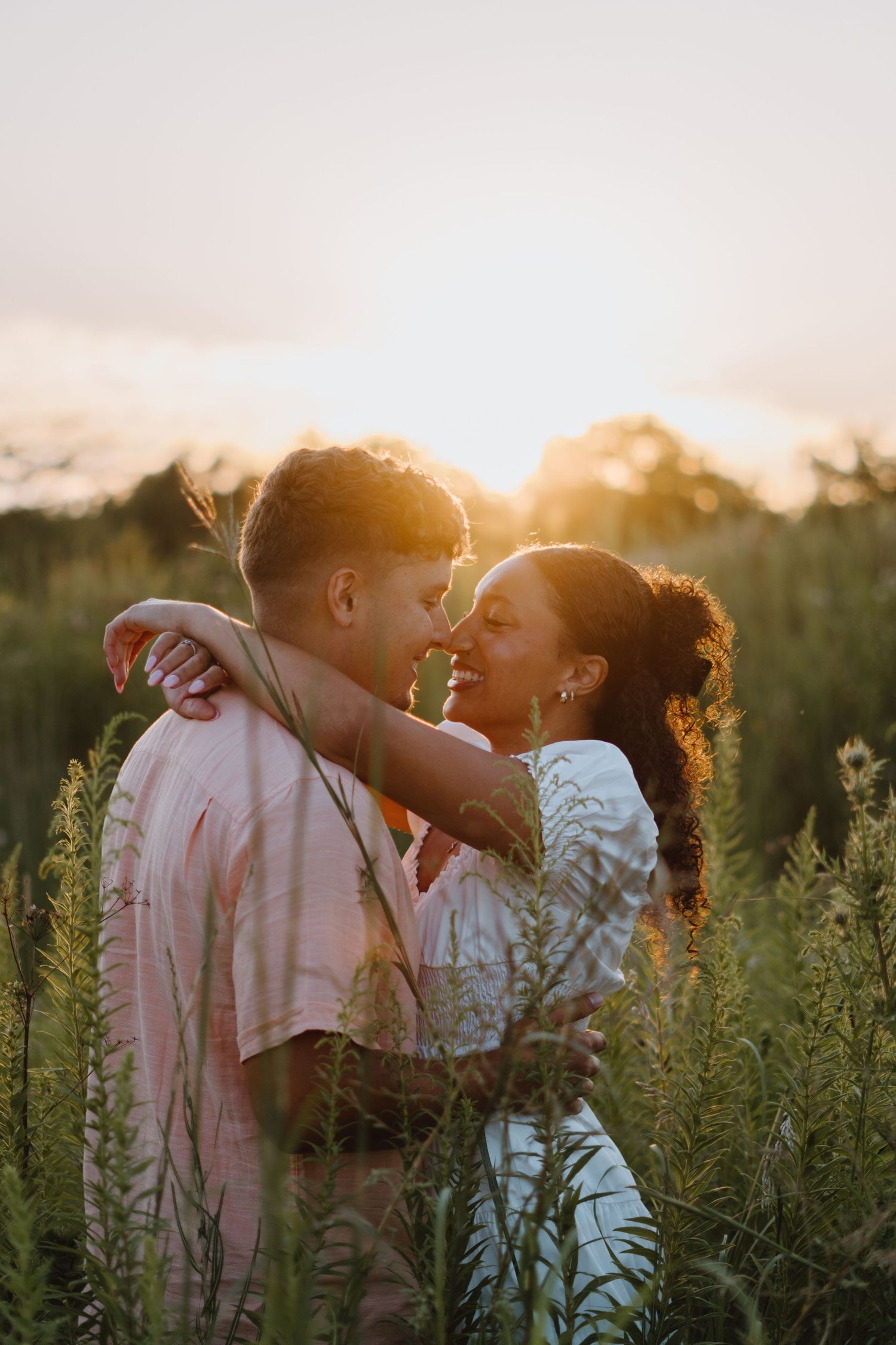 Couple sharing a tender moment nose-to-nose surrounded by tall green wildflowers during a warm sunset.