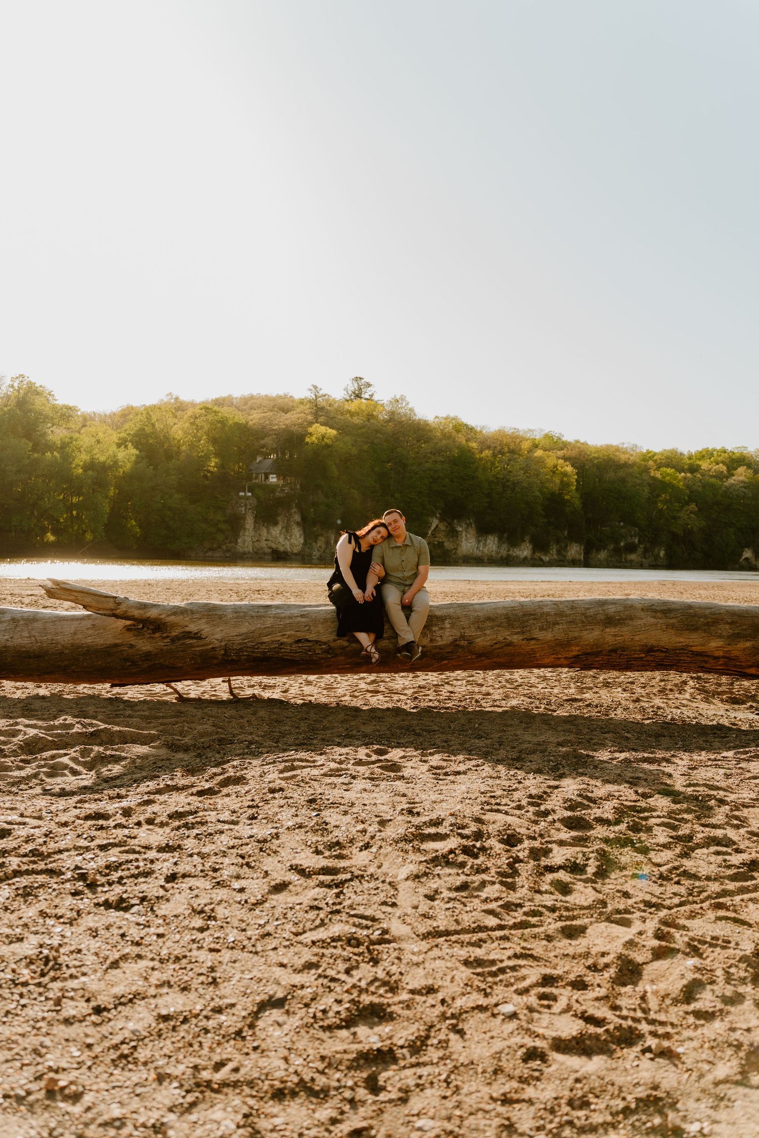 Couple sitting closely on a driftwood log on a sandy beach at golden hour with lush green trees in background.