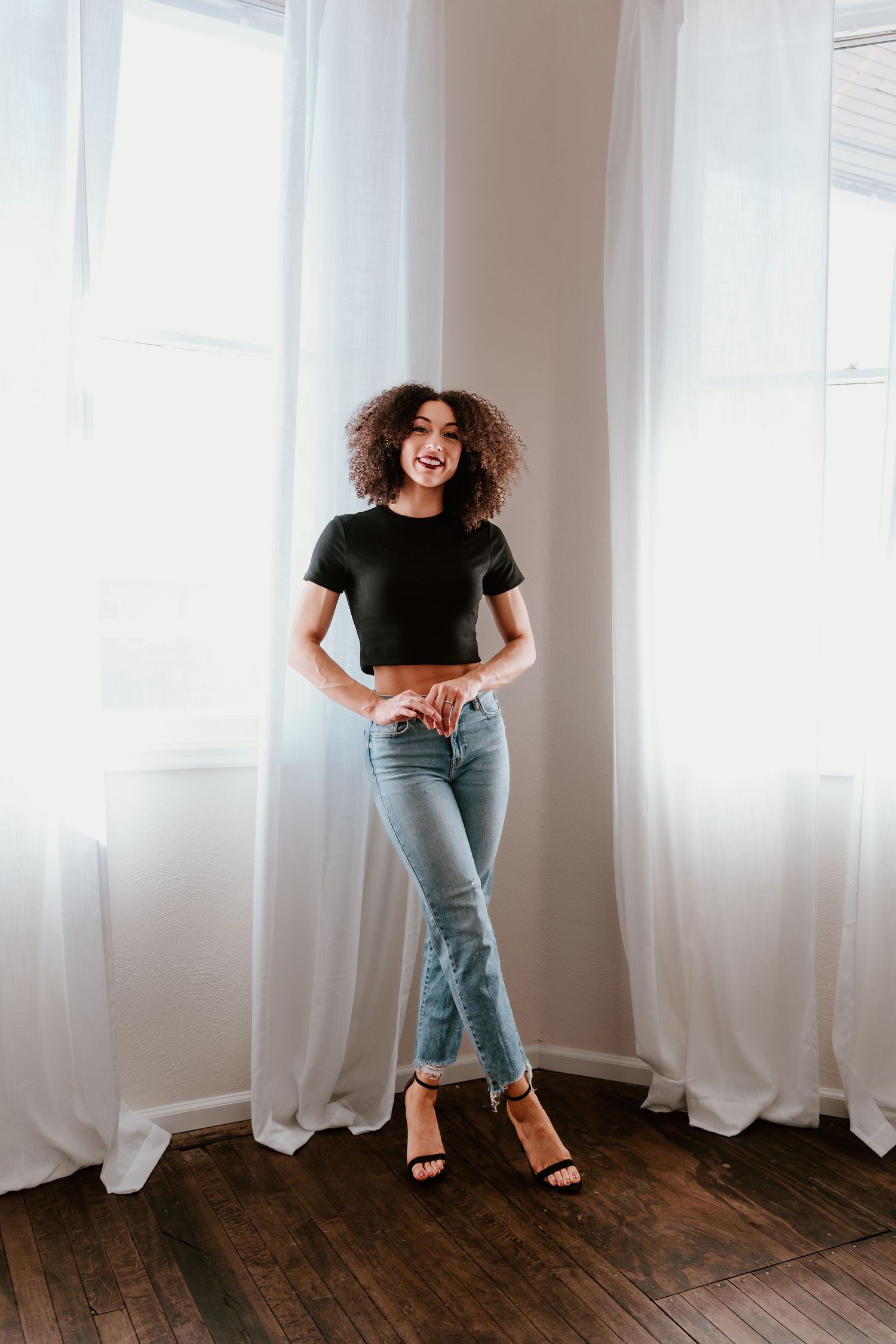 A young woman with curly hair wearing a black crop top, light blue jeans, and heeled sandals stands smiling by bright windows.