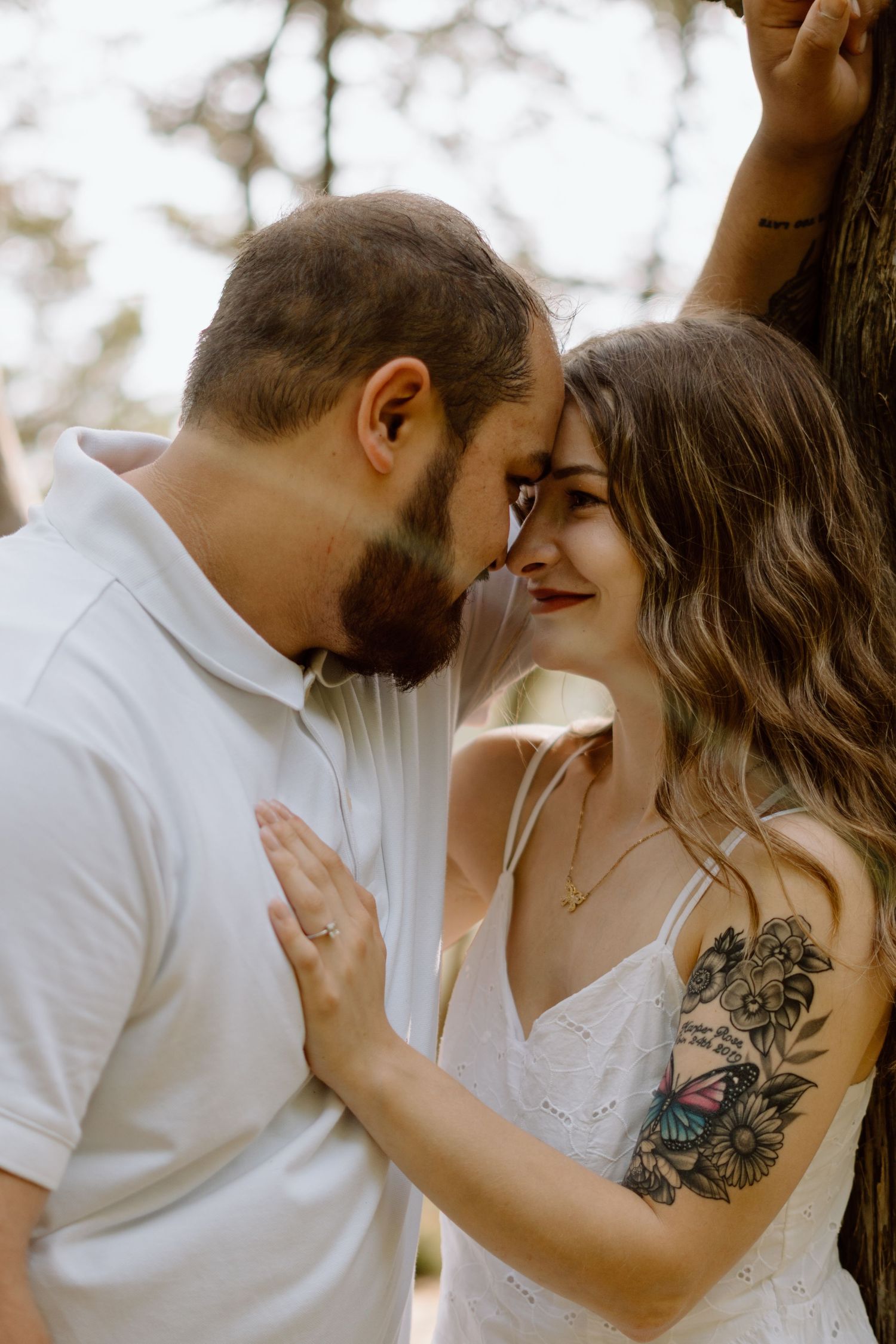 A couple lean in together tenderly against a tree outdoors.