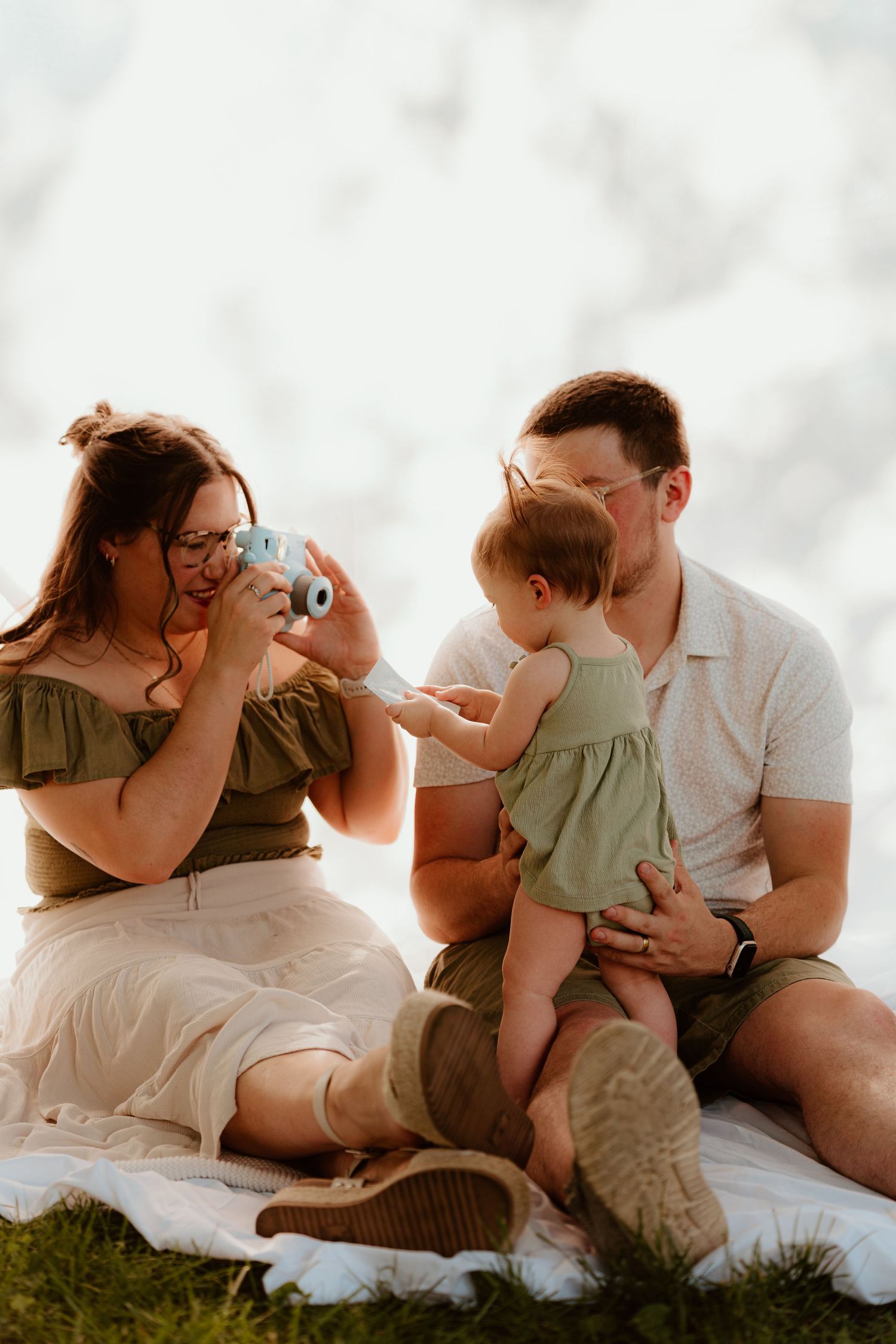 A family sits on a blanket outdoors, mom photographs baby with instant camera while dad holds the infant.