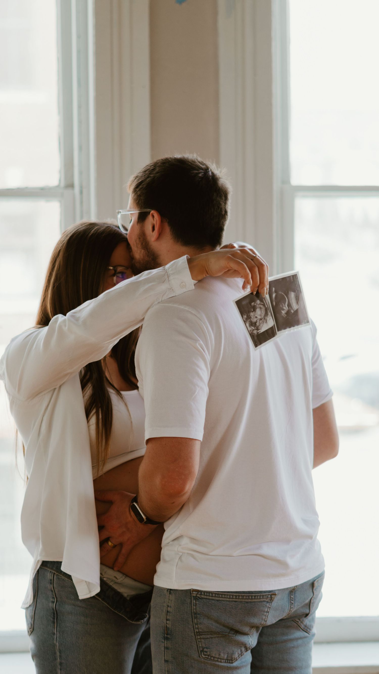 Couple shares a tender kiss while holding ultrasound photos, announcing their pregnancy near a bright window.