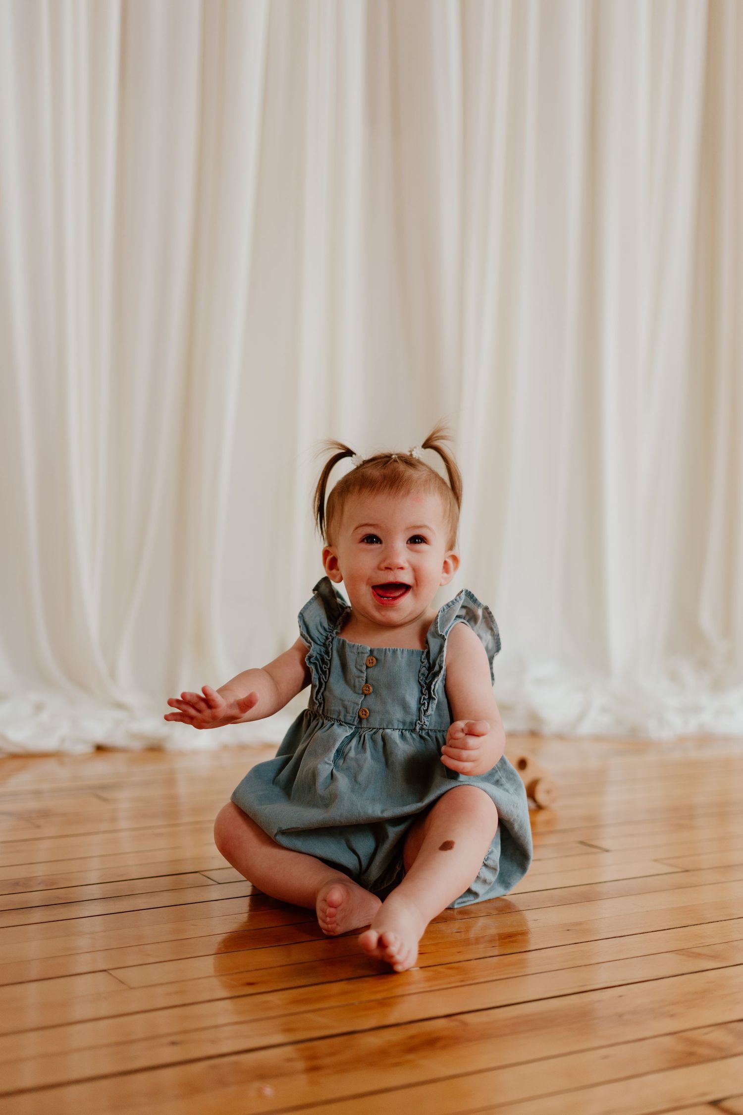 Happy baby girl in blue dress with pigtails sitting on hardwood floor, laughing joyfully in front of white curtains.