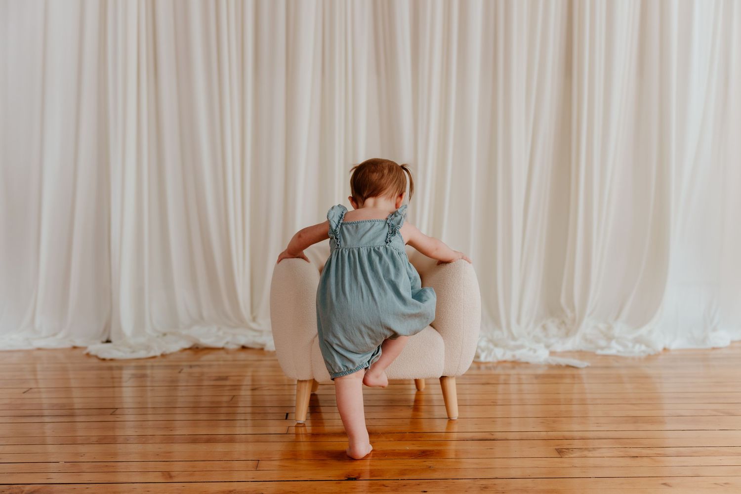 Baby girl in blue romper standing on small white chair against white curtain backdrop on hardwood floor.