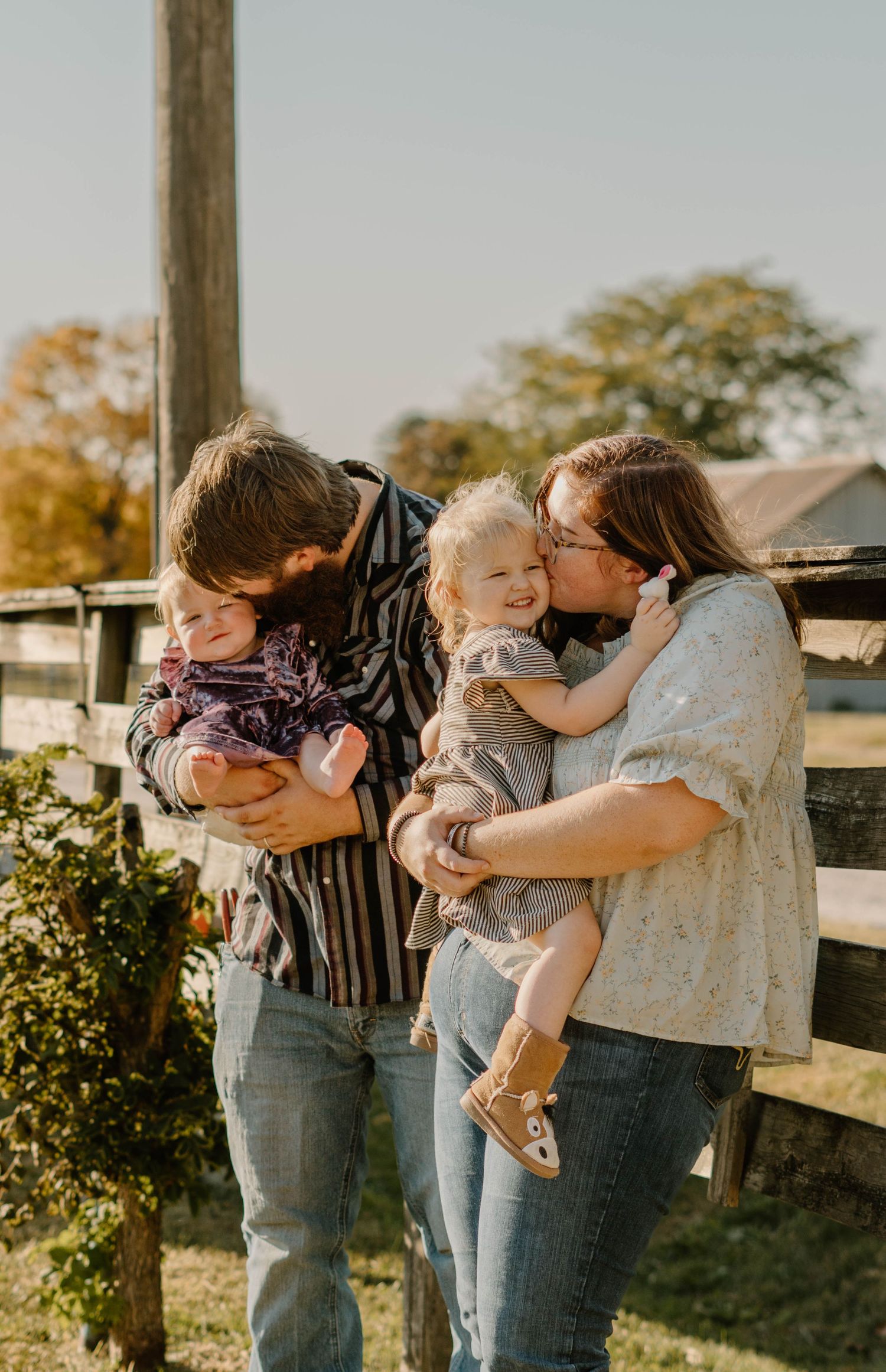 Parents lovingly kissing their two young children near a rustic wooden fence on a bright autumn day.