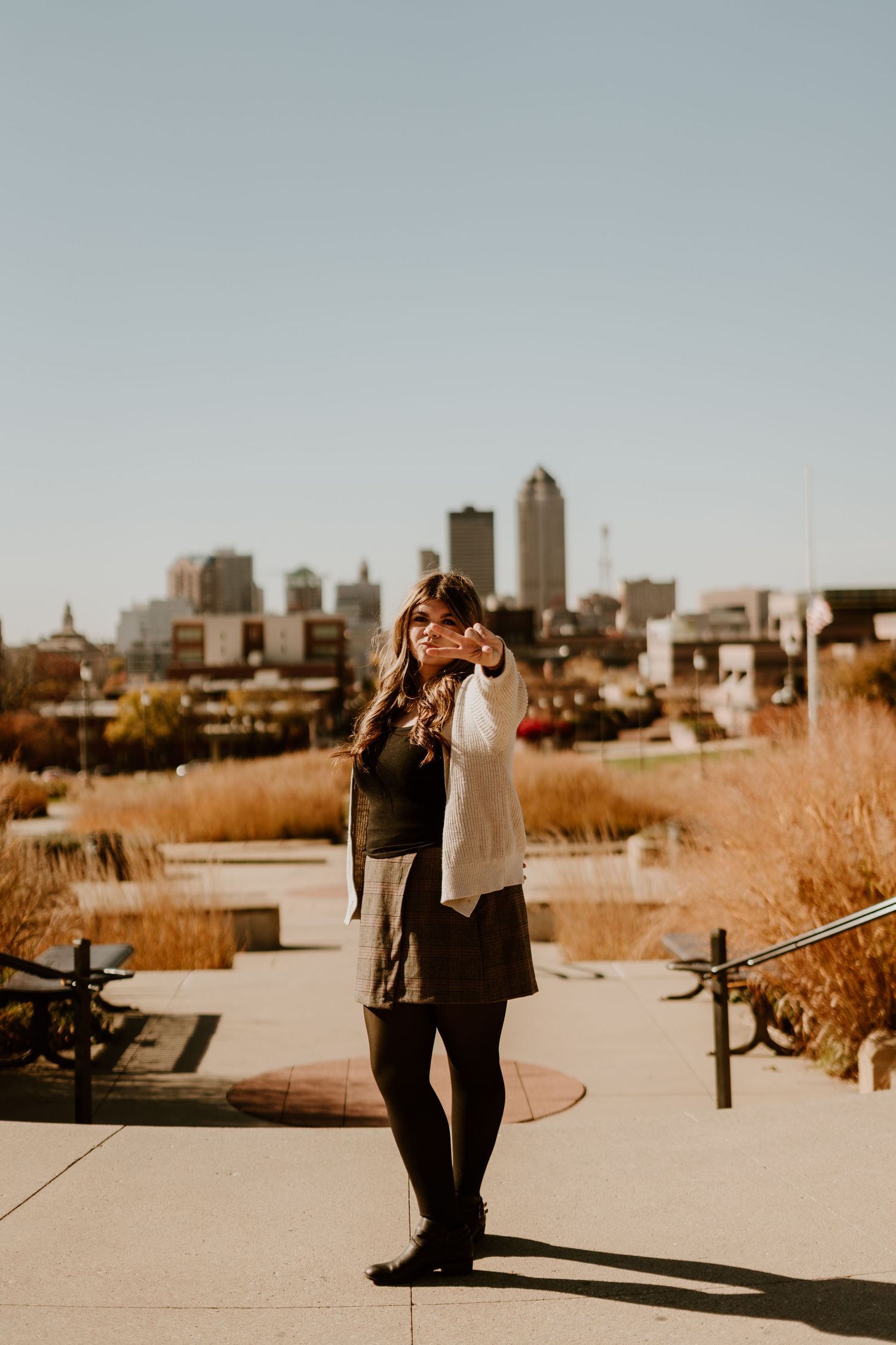 Woman posing outdoors with city skyline and golden grasses in background on a sunny day.