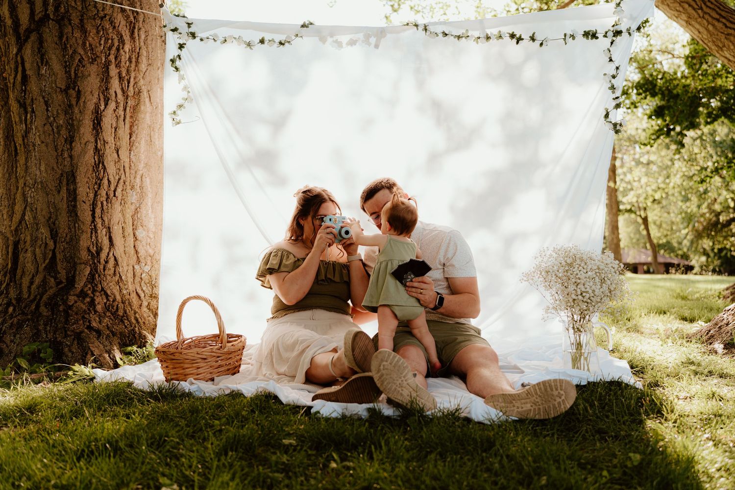 Family enjoying a sunny outdoor picnic under a white canopy backdrop, sitting on a blanket with a baby near a large tree.
