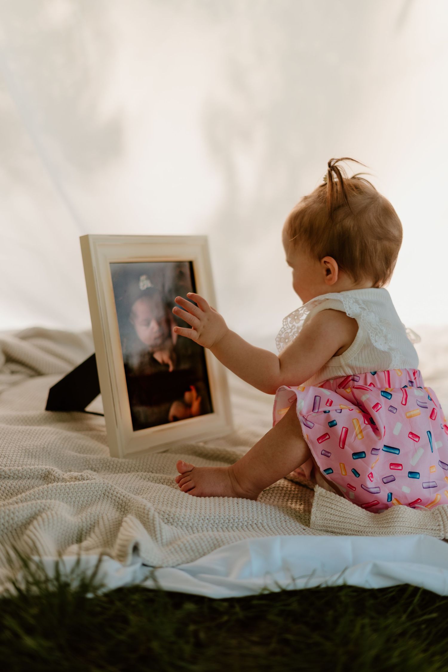 Baby girl in pink dress touching a framed photo while sitting on a blanket outdoors.
