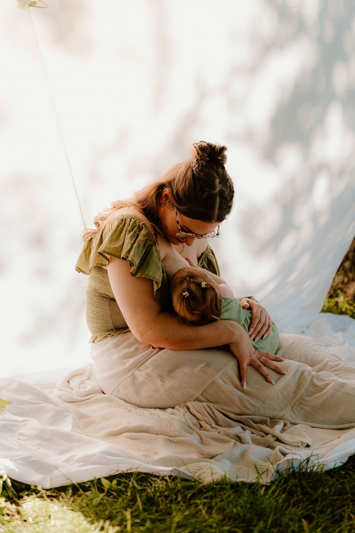 Mother tenderly holds and nurses her baby while sitting on a blanket outdoors in soft natural light.