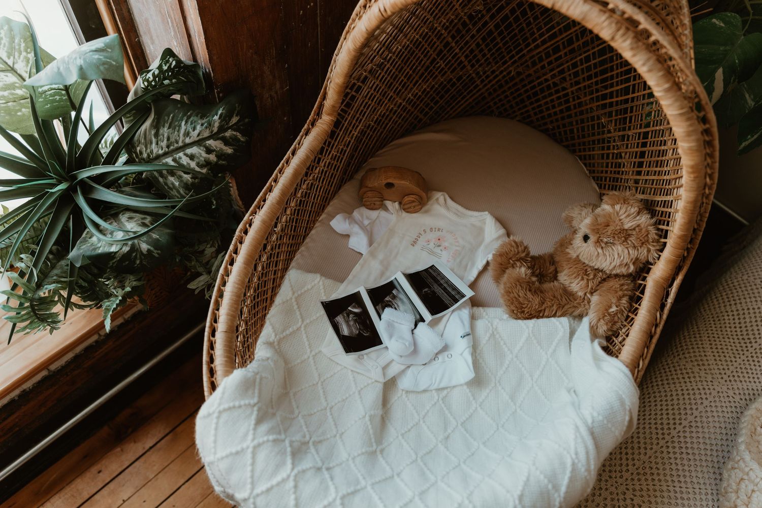 Wicker bassinet with ultrasound photos, baby clothes, and teddy bear on white blanket near window with plants.