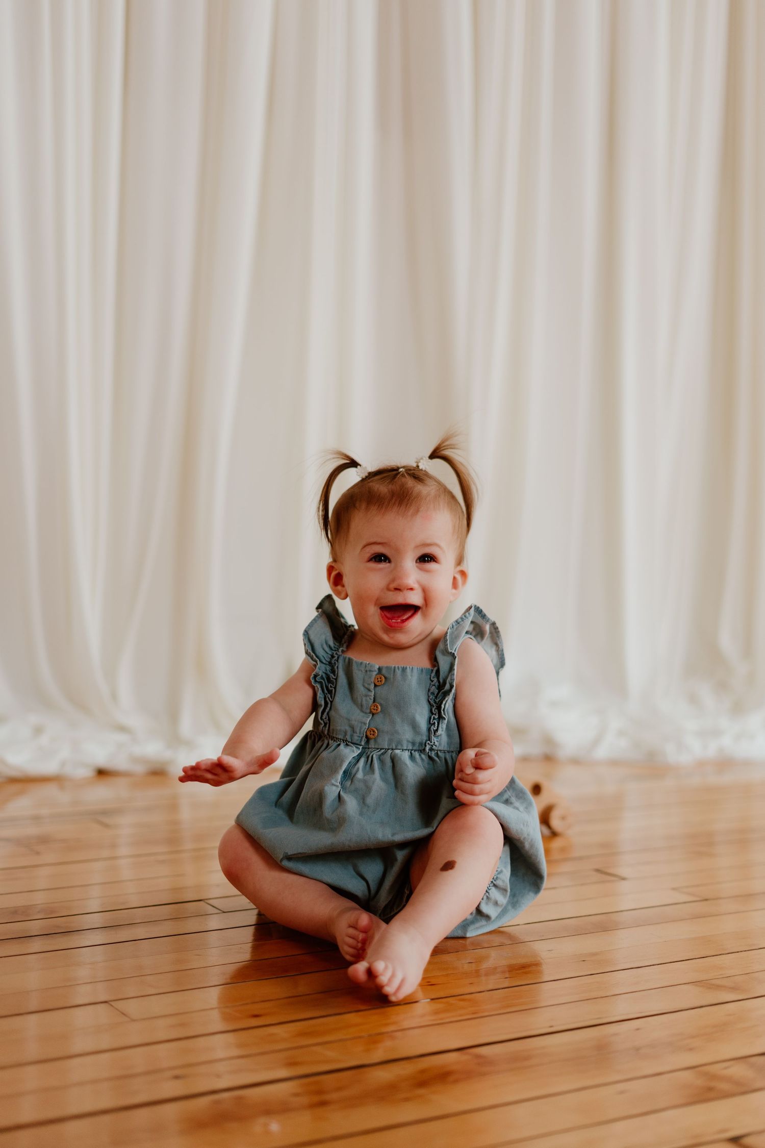 Laughing baby girl in blue dress with pigtails sitting on hardwood floor against white curtain backdrop.