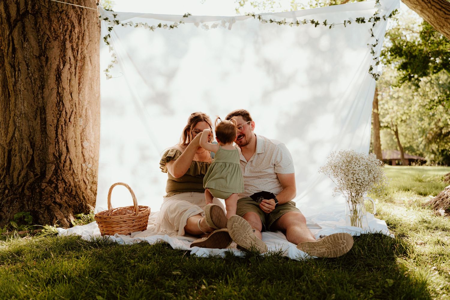 Family of three sitting on white blanket under tree with picnic basket and floral backdrop outdoors.