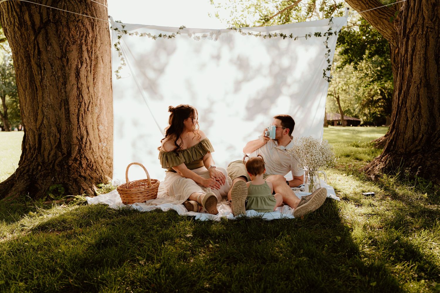Family enjoying a sunny outdoor picnic on a blanket between two trees with a white backdrop and string lights.