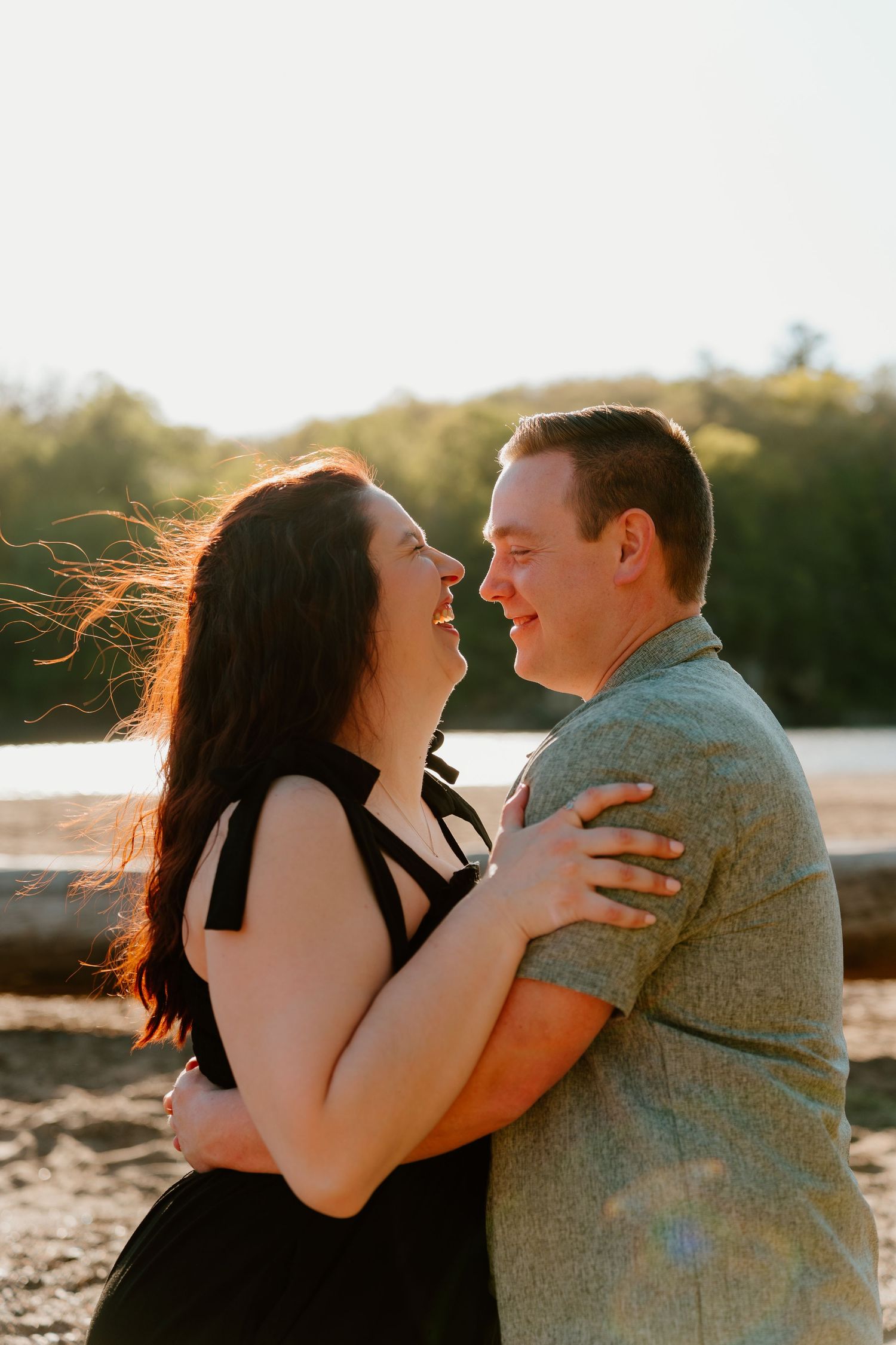 Couple embracing and smiling at each other on a beach at golden hour, woman in black dress, man in gray shirt.