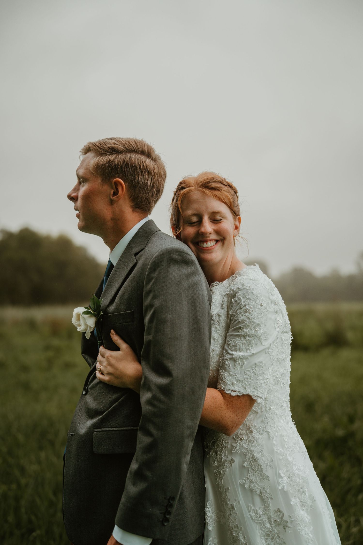 A smiling bride in a lace dress hugs a groom in a gray suit from behind in a green field.