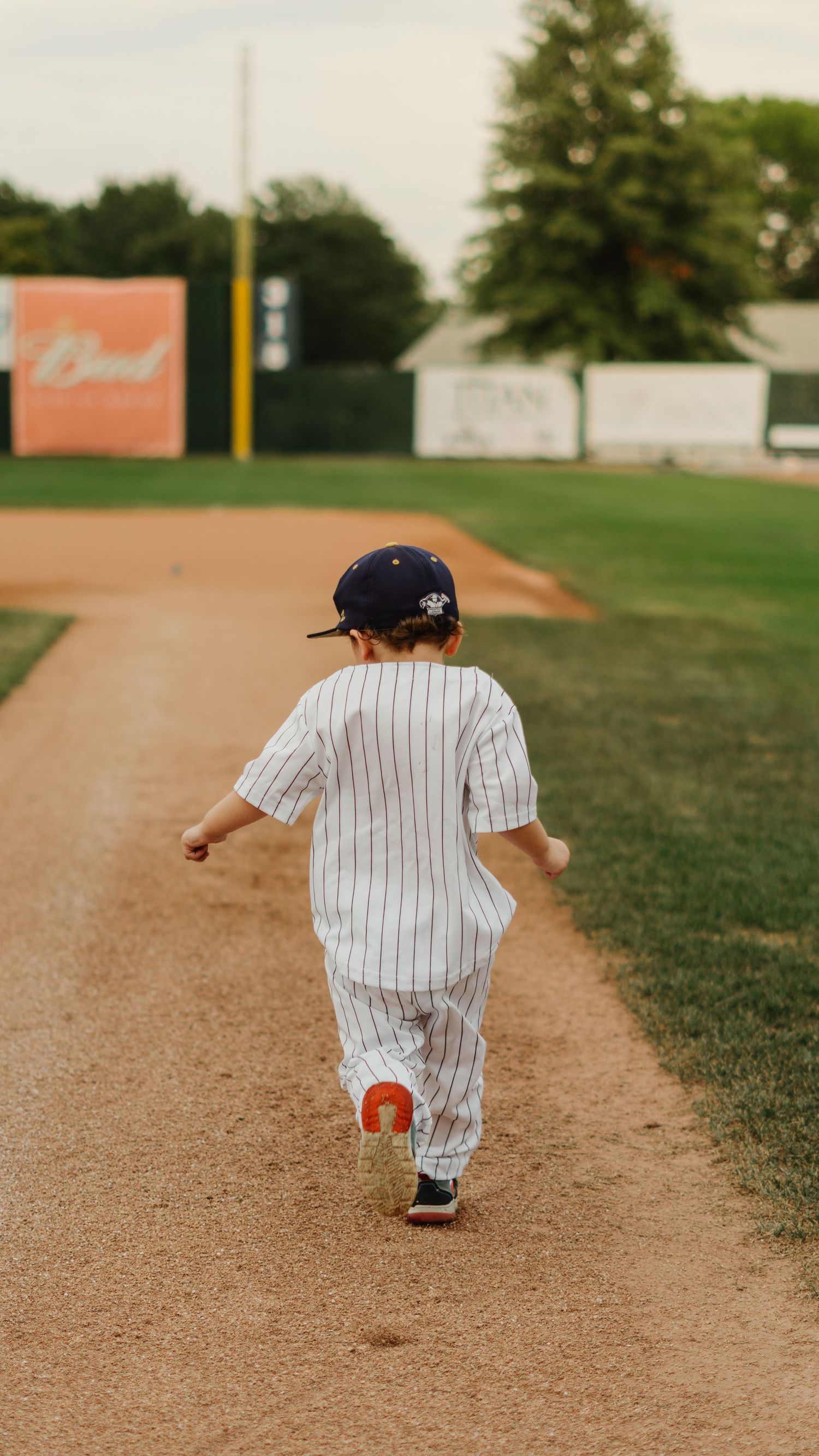A toddler in a pinstripe baseball uniform and navy cap runs along the baseline of a baseball field.