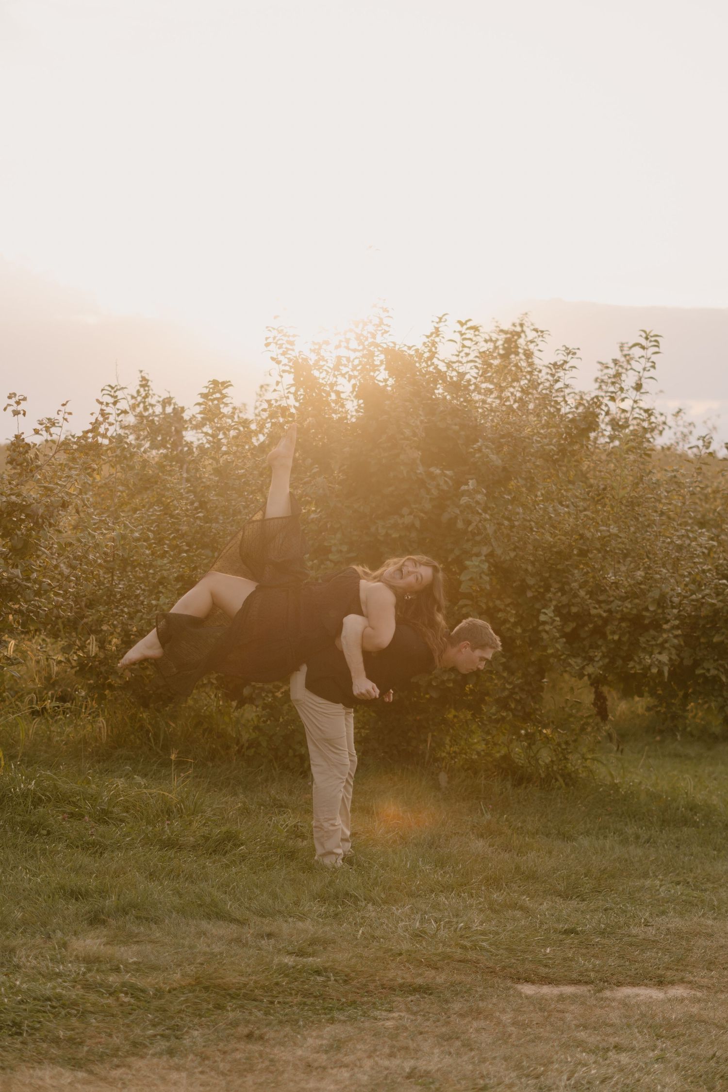 Couple playing in golden sunset light in an orchard, man carrying woman on his back among lush green trees.