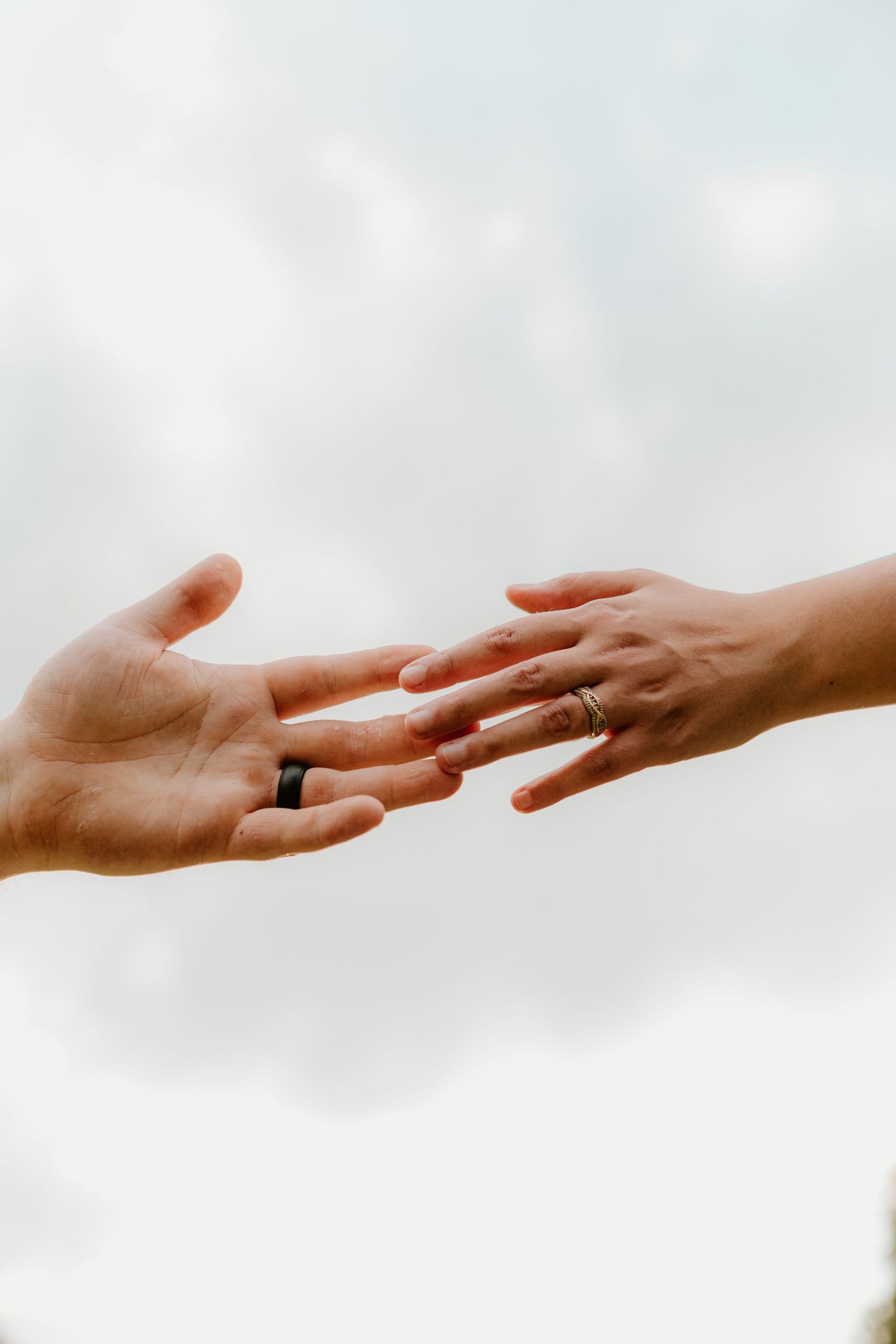 Two hands reaching toward each other, both wearing wedding bands, against a soft cloudy sky background.